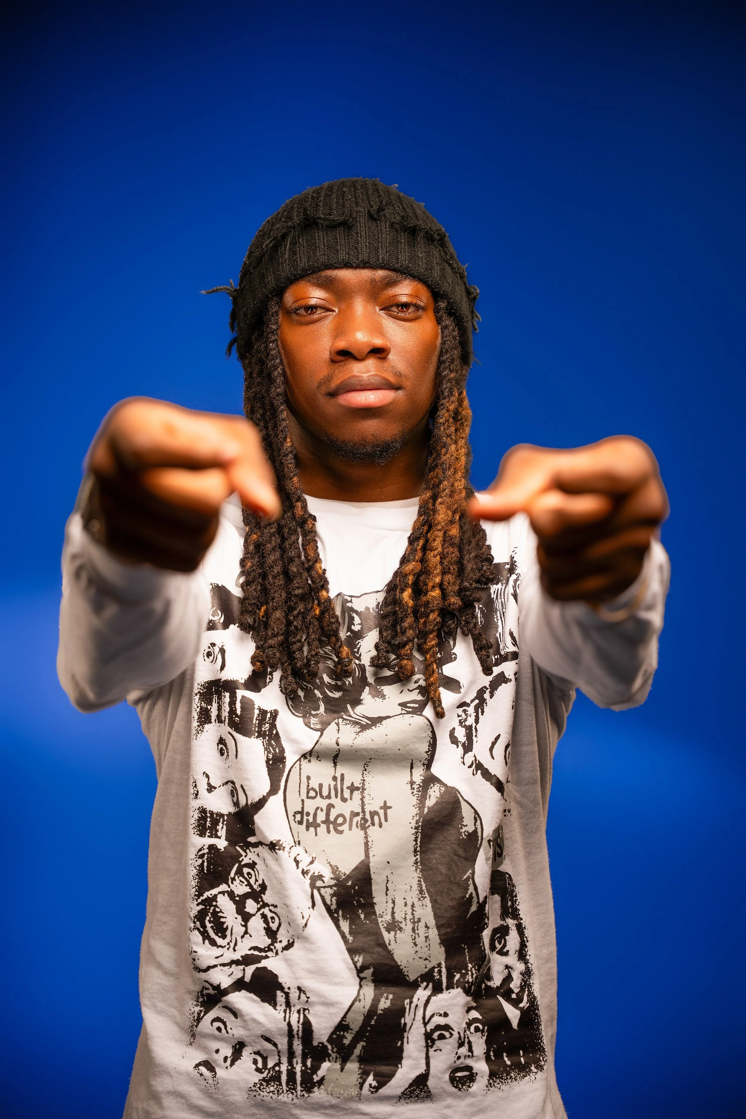 A young man with dreadlocks and a black beanie is pointing towards the camera against a vibrant blue background.