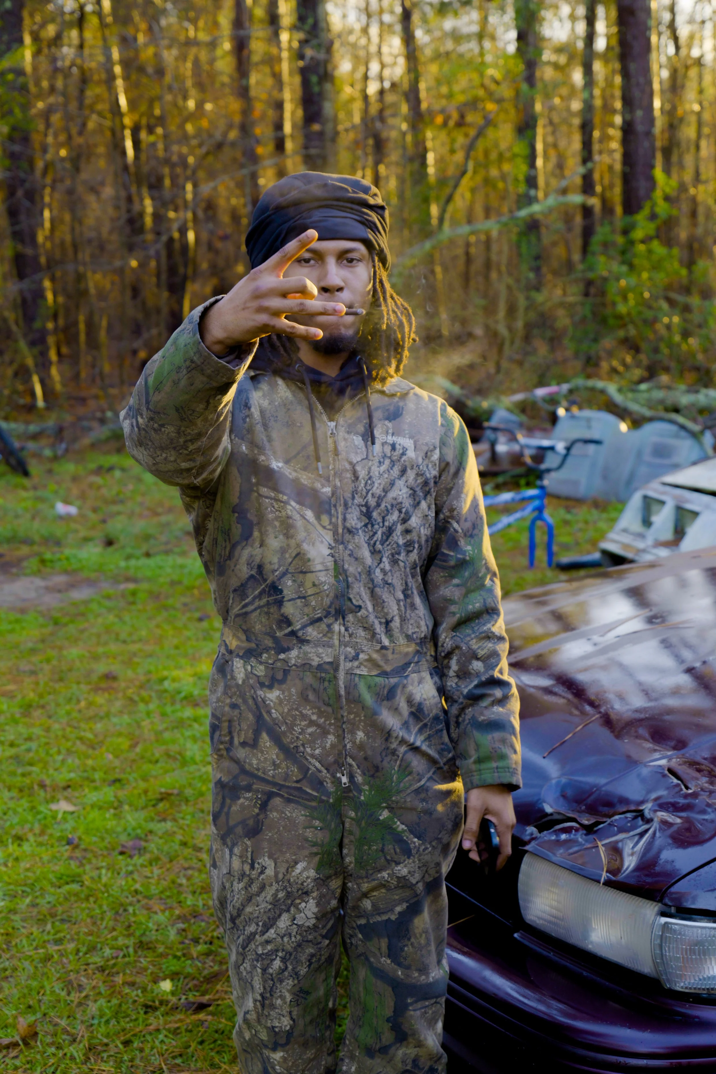 A young man with dreadlocks wearing camouflage clothing standing outdoors near a damaged car in a wooded area during daytime.