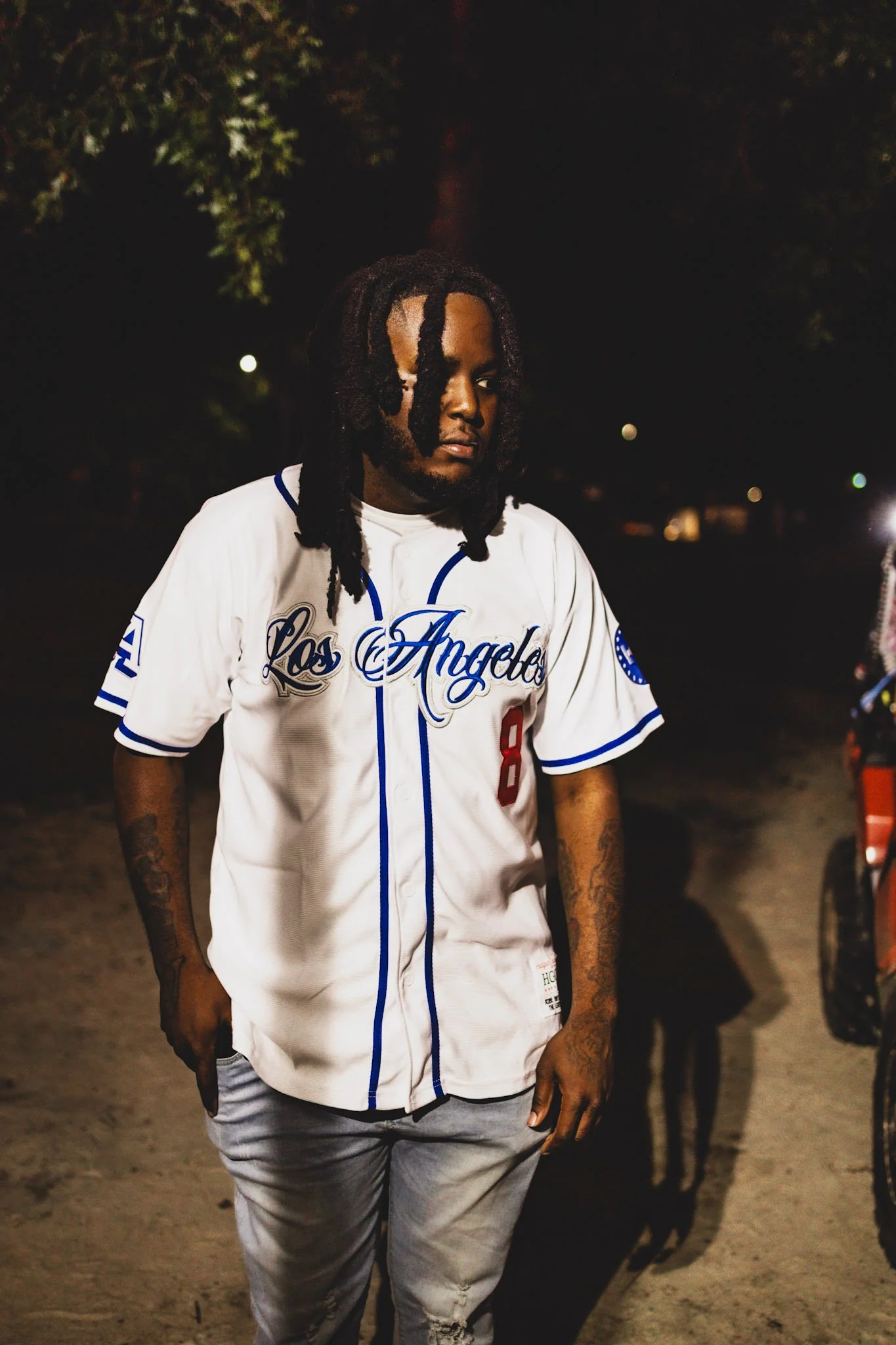 A young man with dreadlocks wearing a white Los Angeles sports jersey, standing outdoors at night.