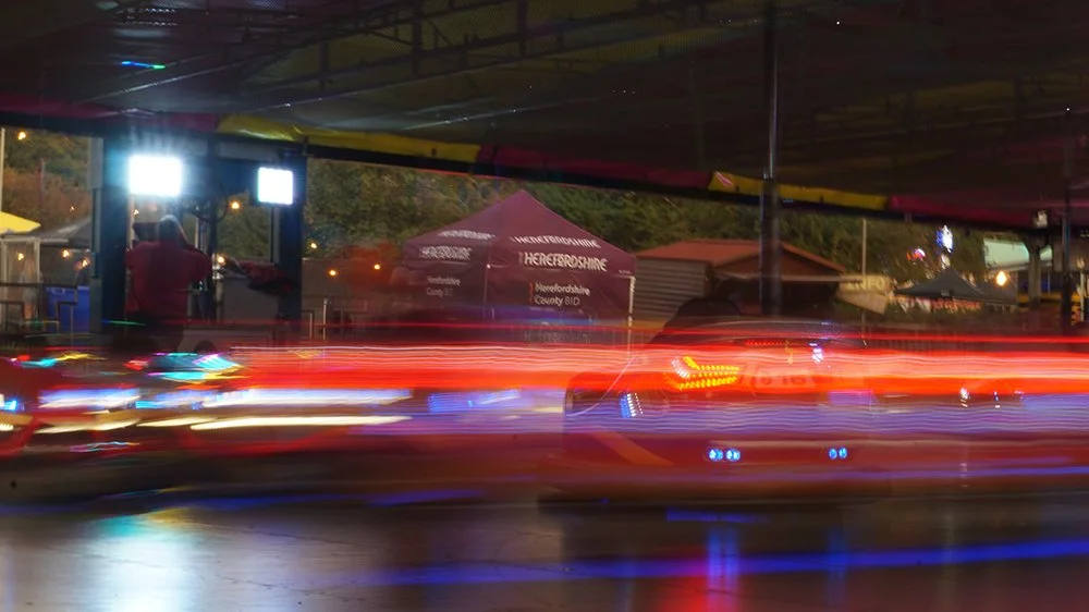 Blurred image of a night scene with streaks of red, white, and blue light from moving cars. In the background, a tent with Pennine Moorland signage, a person in a red shirt, and some structures are visible.