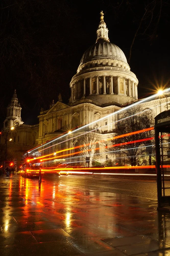 Night view of St. Paul's Cathedral in London with light trails from passing vehicles, wet pavement reflecting city lights, and a traditional red telephone booth in the foreground.