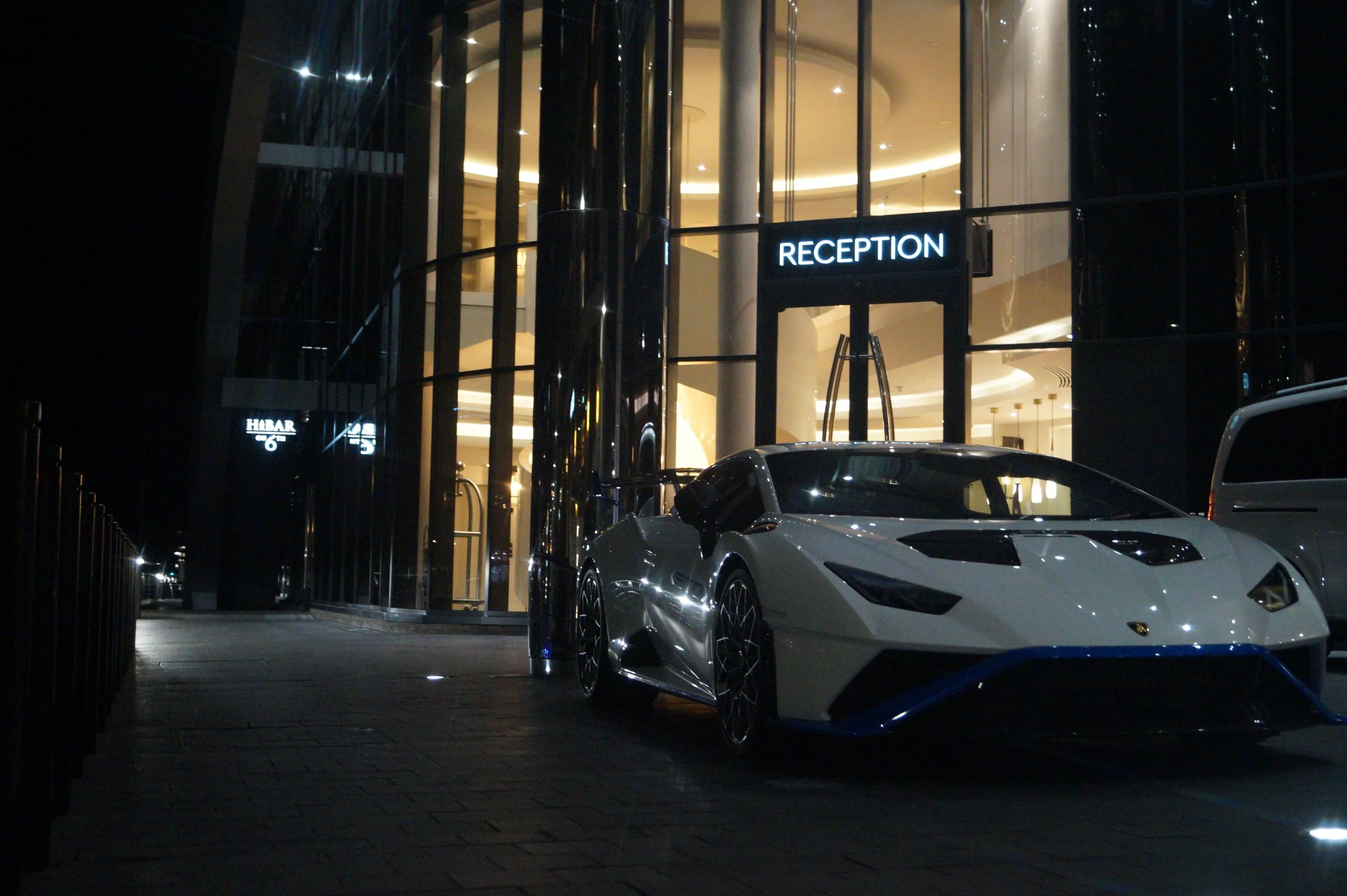 A white luxury sports car parked outside a modern building with illuminated 'RECEPTION' sign at night.