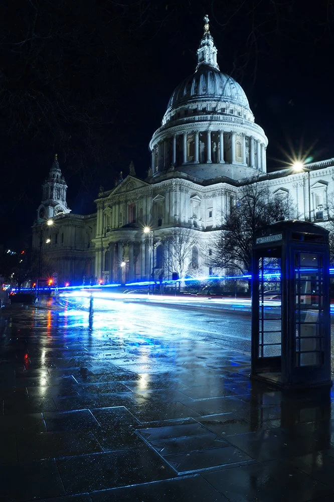 Nighttime view of St. Paul's Cathedral in London, illuminated with white lights, with a wet street and a police phone booth in the foreground.
