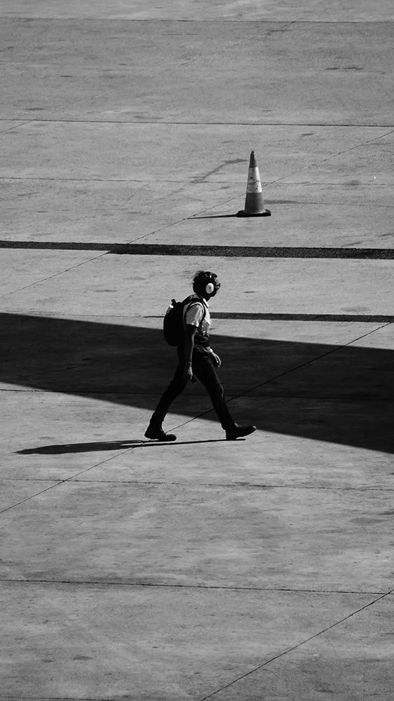 A person walking on a concrete surface with a backpack, wearing headphones, and casting a shadow with a traffic cone in the background.