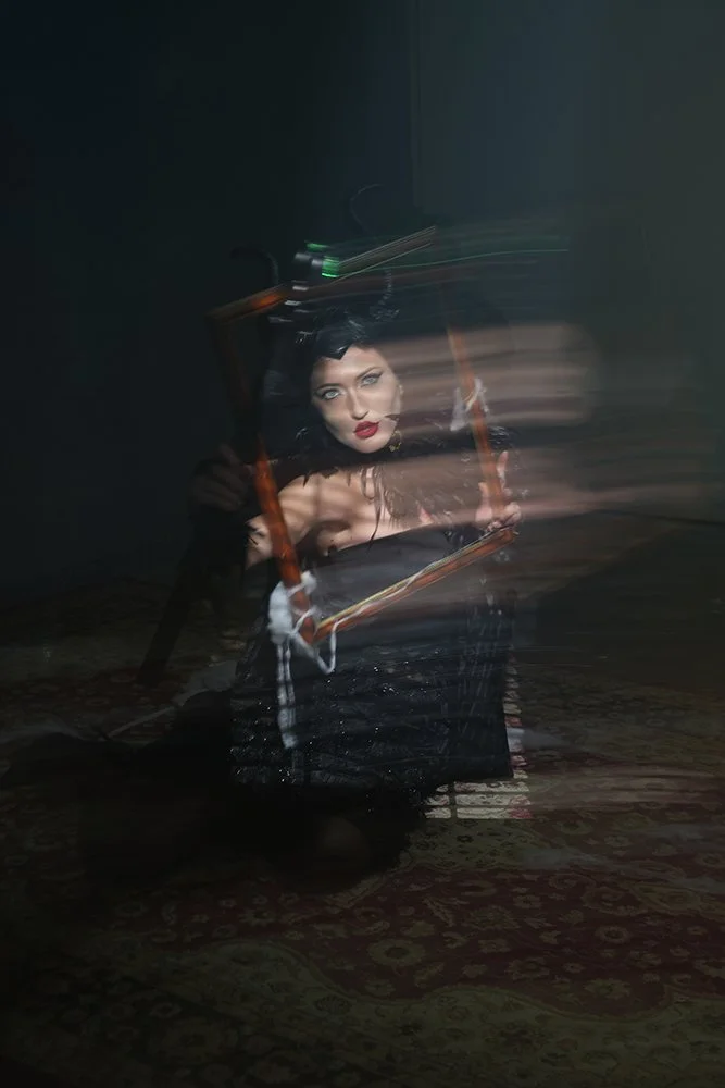 A woman with dark hair, dressed in black, holding a frame or mirror, with a significant motion blur effect creating streaks of light and motion around her.