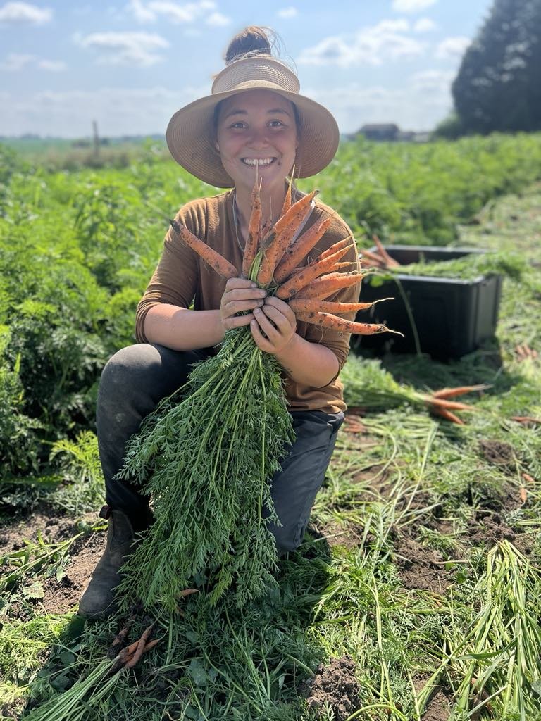 A smiling woman in a wide-brimmed hat kneeling in a garden holding a bunch of freshly picked carrots with green tops.