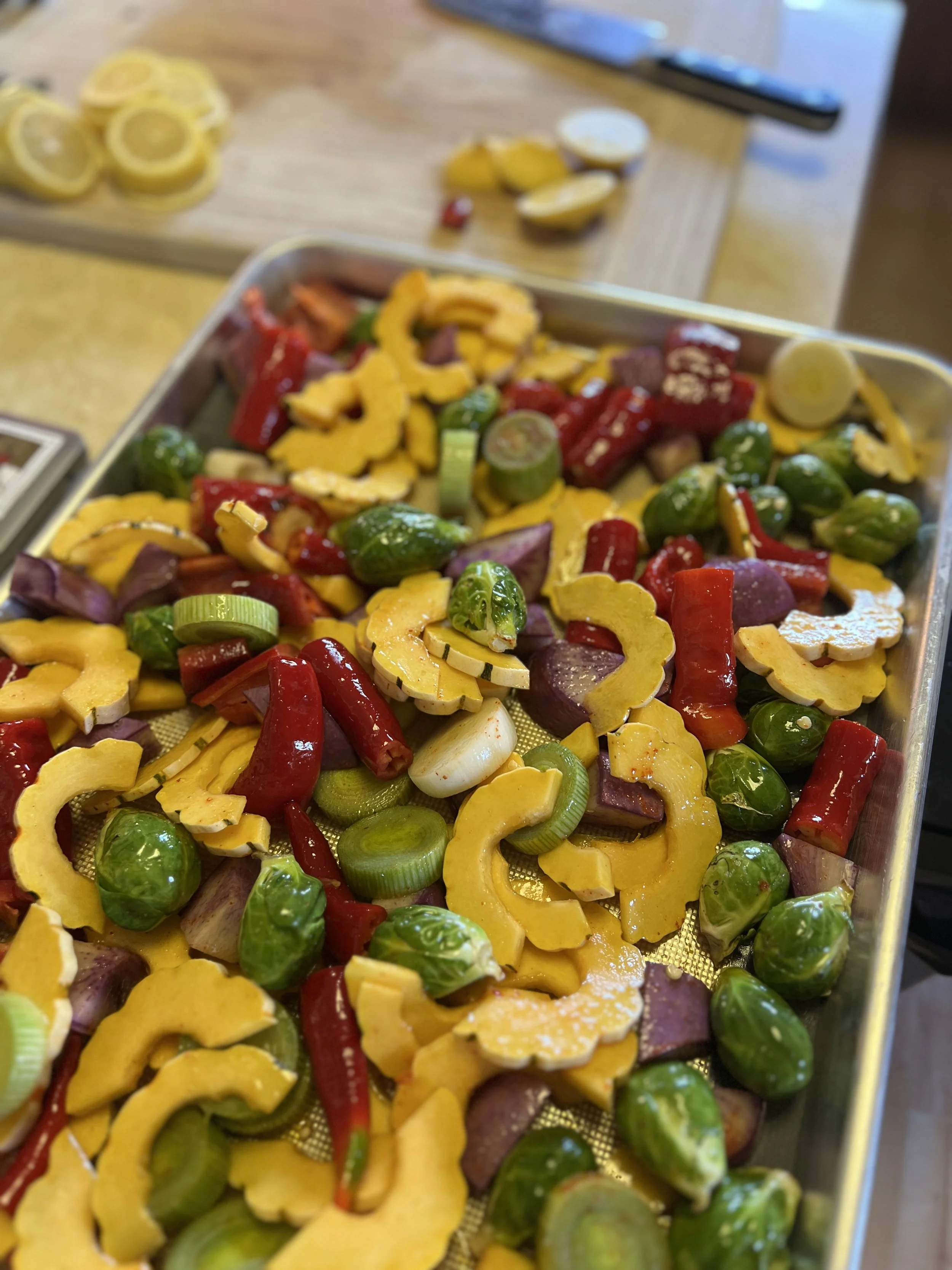 Baking sheet of mixed roasted vegetables including yellow squash, red bell peppers, green Brussels sprouts, purple onions, and celery, with a cutting board of lemon slices and a knife in the background.