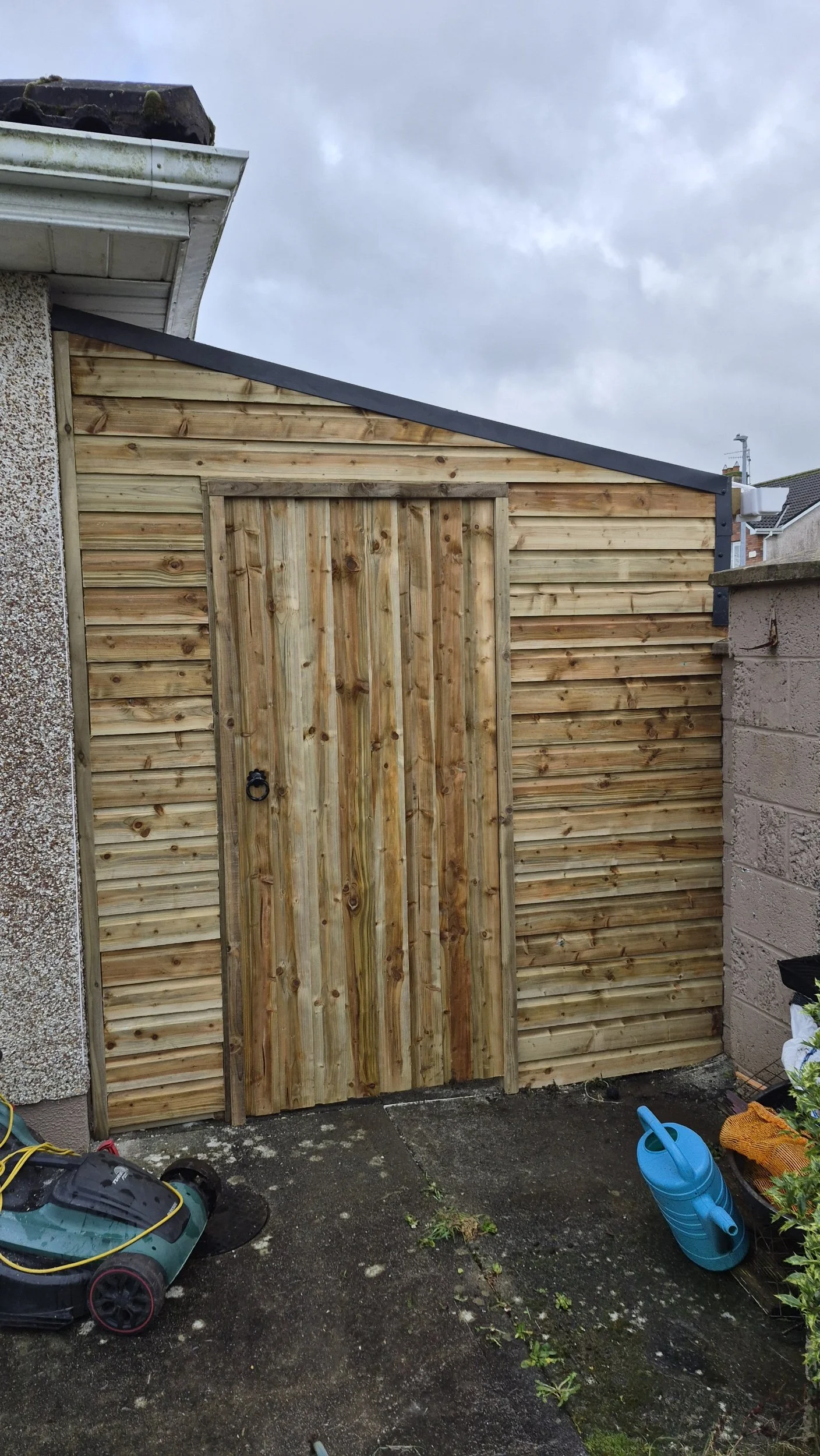A new wooden garden shed with a door, located outdoors on a paved patio area, with gardening tools and supplies nearby, under a cloudy sky.