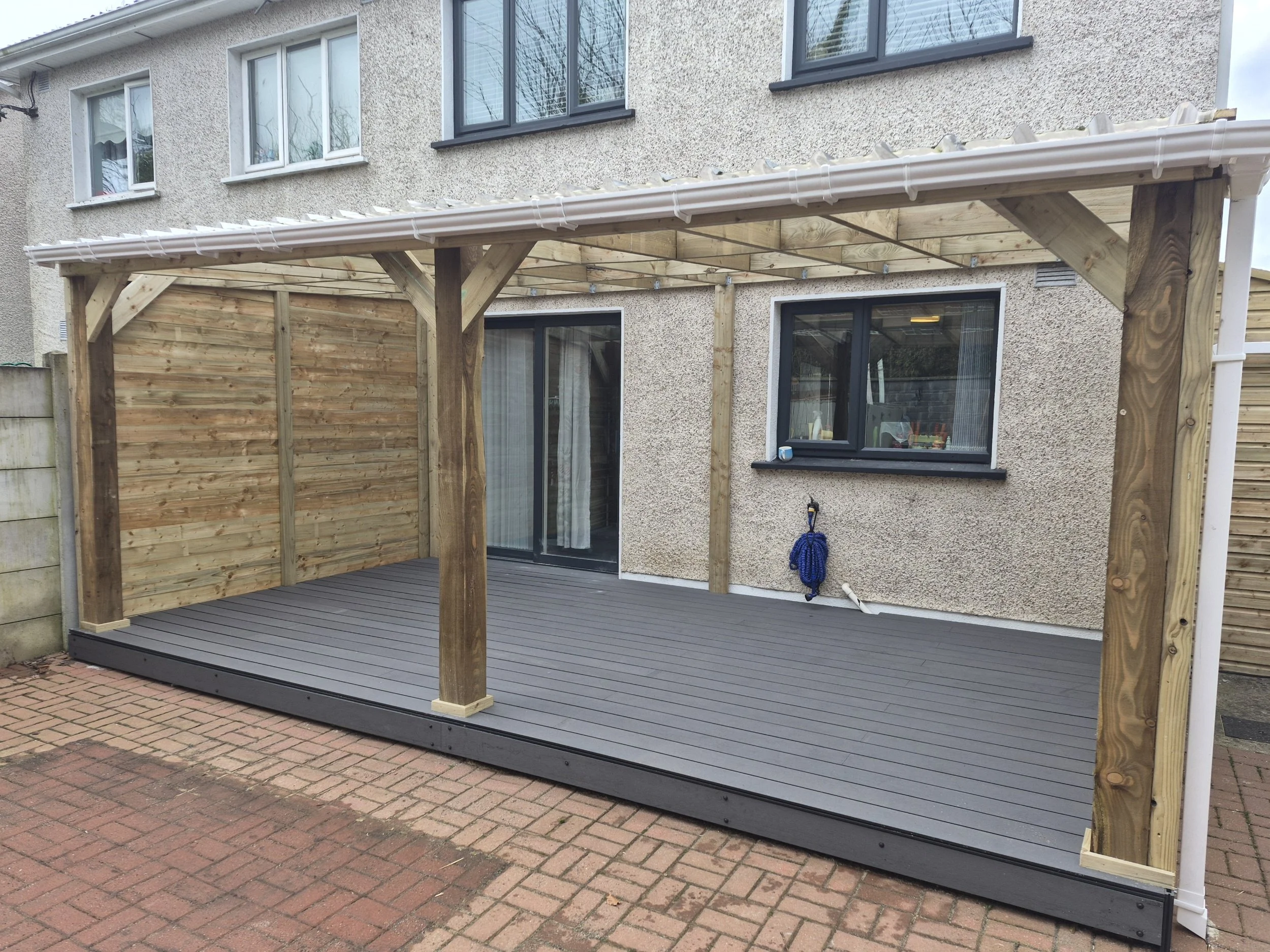 A wooden deck with a deck cover structure attached to the back of a two-story house. The house has beige textured walls, black window frames, and a sliding glass door. A blue garden hose hangs from a hook on the wall. The deck is elevated, with a bri
