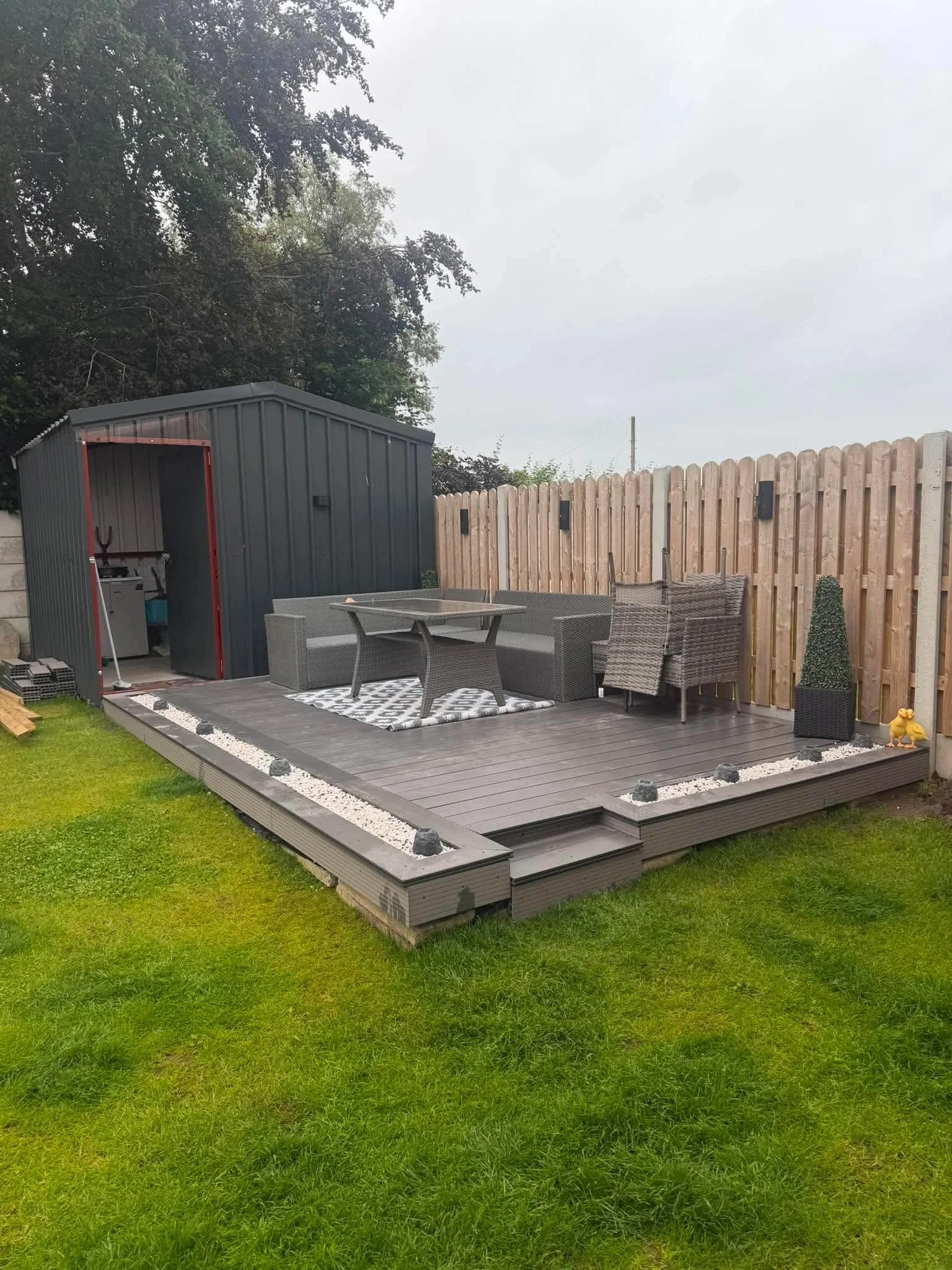 A backyard patio with a grey deck, outdoor furniture, a small grey shed, and a wooden fence, with lush green grass in the foreground.