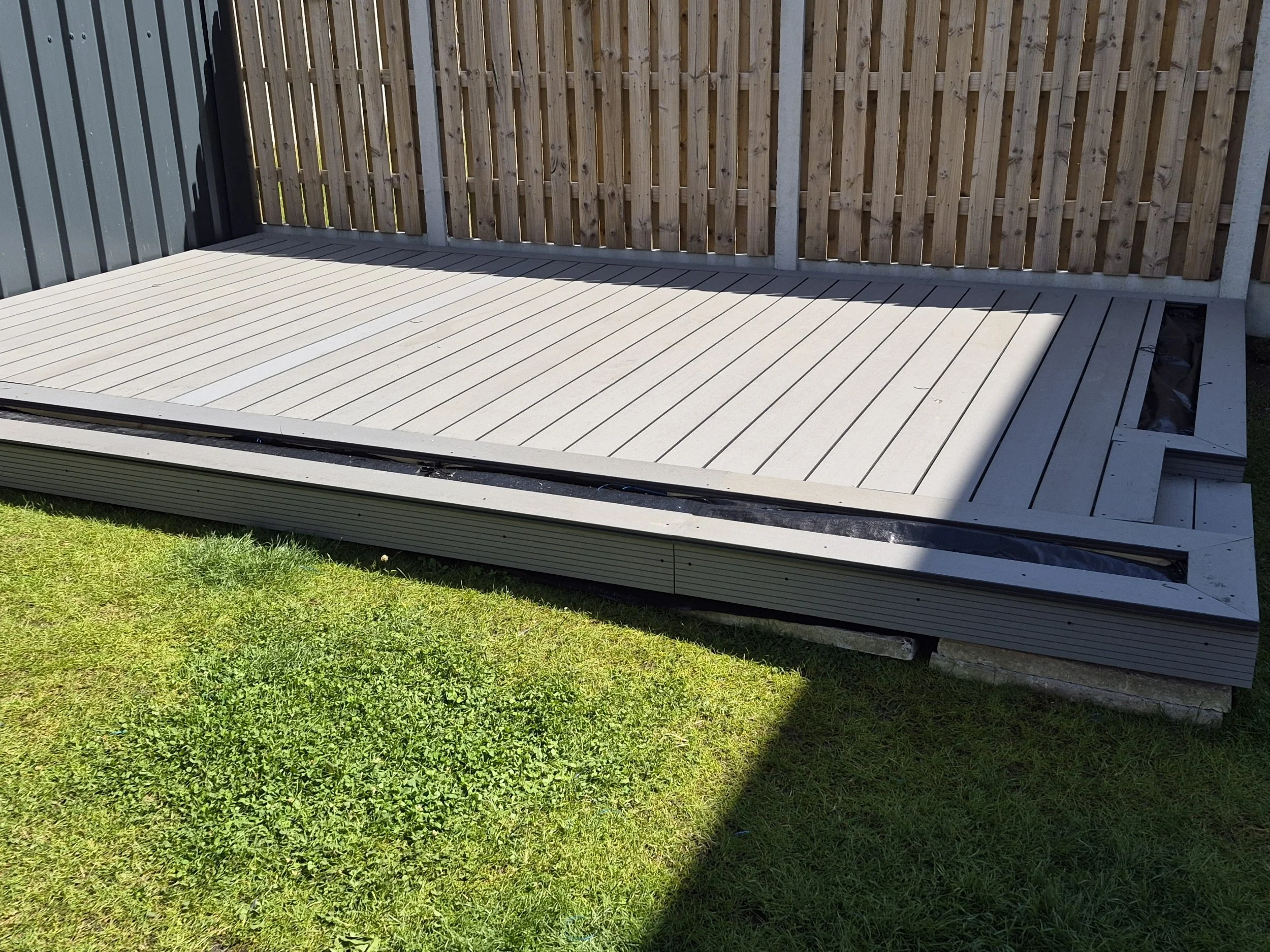 An outdoor wooden deck with grass in the foreground, a wooden fence in the background, and a corner of a metal fence on the left side.