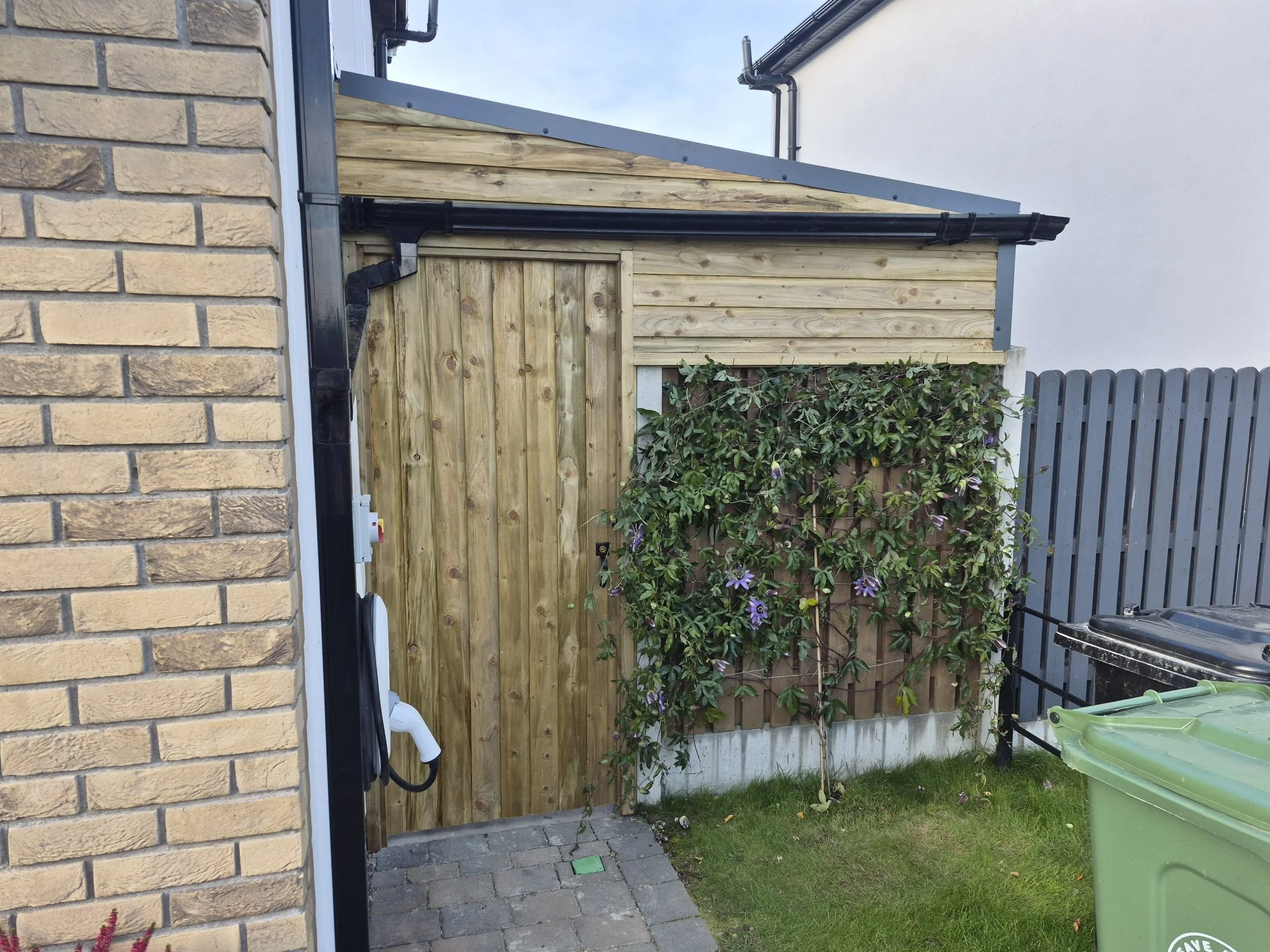 A backyard corner with a wooden fence, a garden with flowering plants, and a gray shed roof. Part of a brick house wall and a gray fence are visible. Some gardening tools and a green plastic container are on the grass.