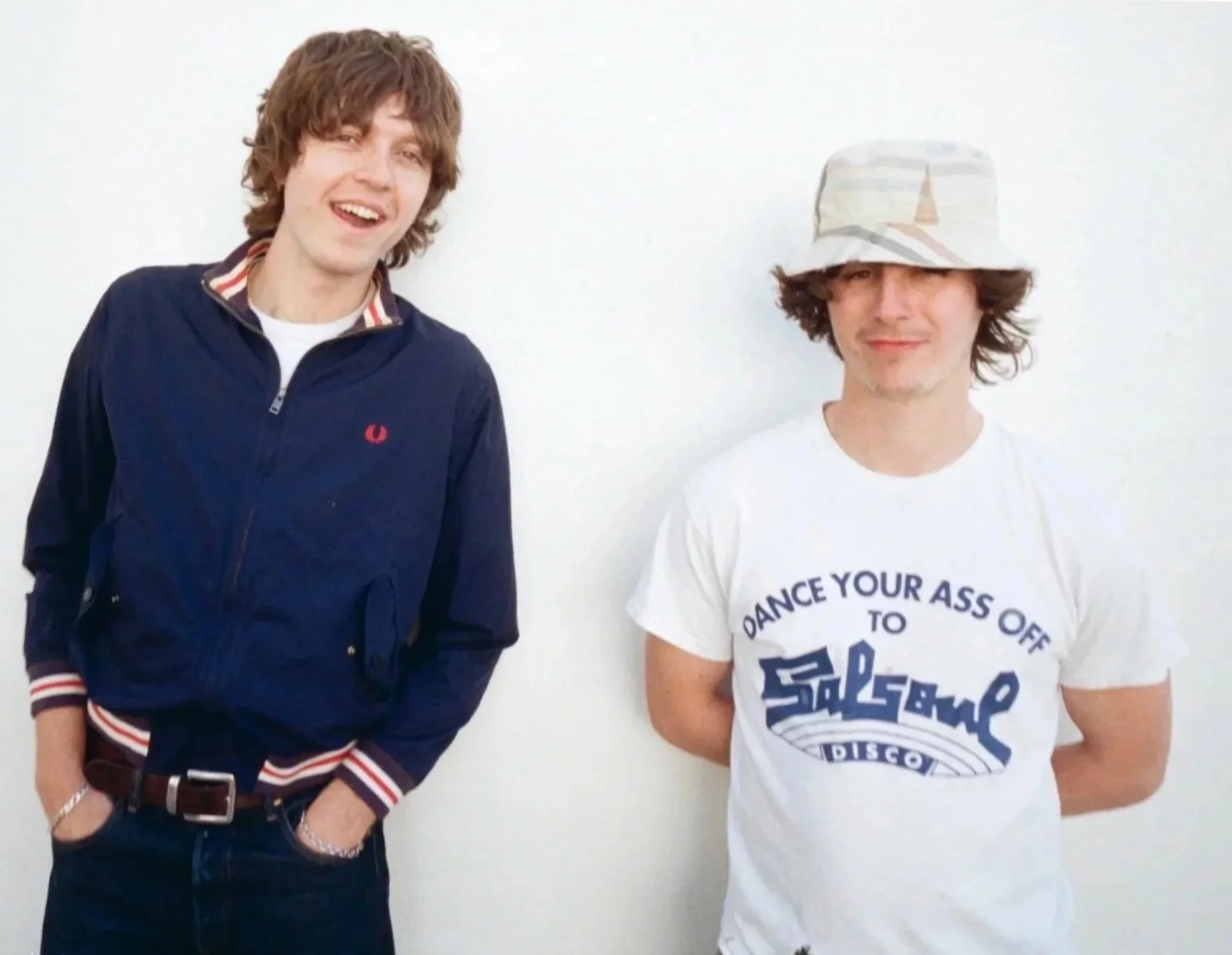 Two young men standing against a plain white wall, smiling. The man on the left is wearing a navy blue jacket with a red and white striped collar and cuffs, dark jeans, and a belt. The man on the right is wearing a white T-shirt with blue text and a graphic, and a light-colored bucket hat with a small sailboat print.