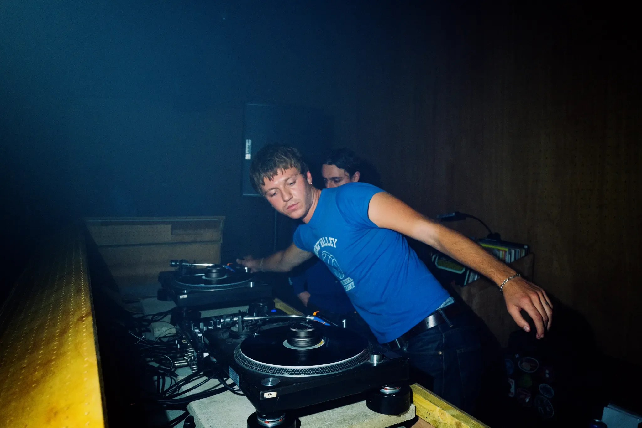 A young man DJing at a music event, standing behind a DJ controller with headphones in his hand, in a dimly lit room with a wooden wall behind him.
