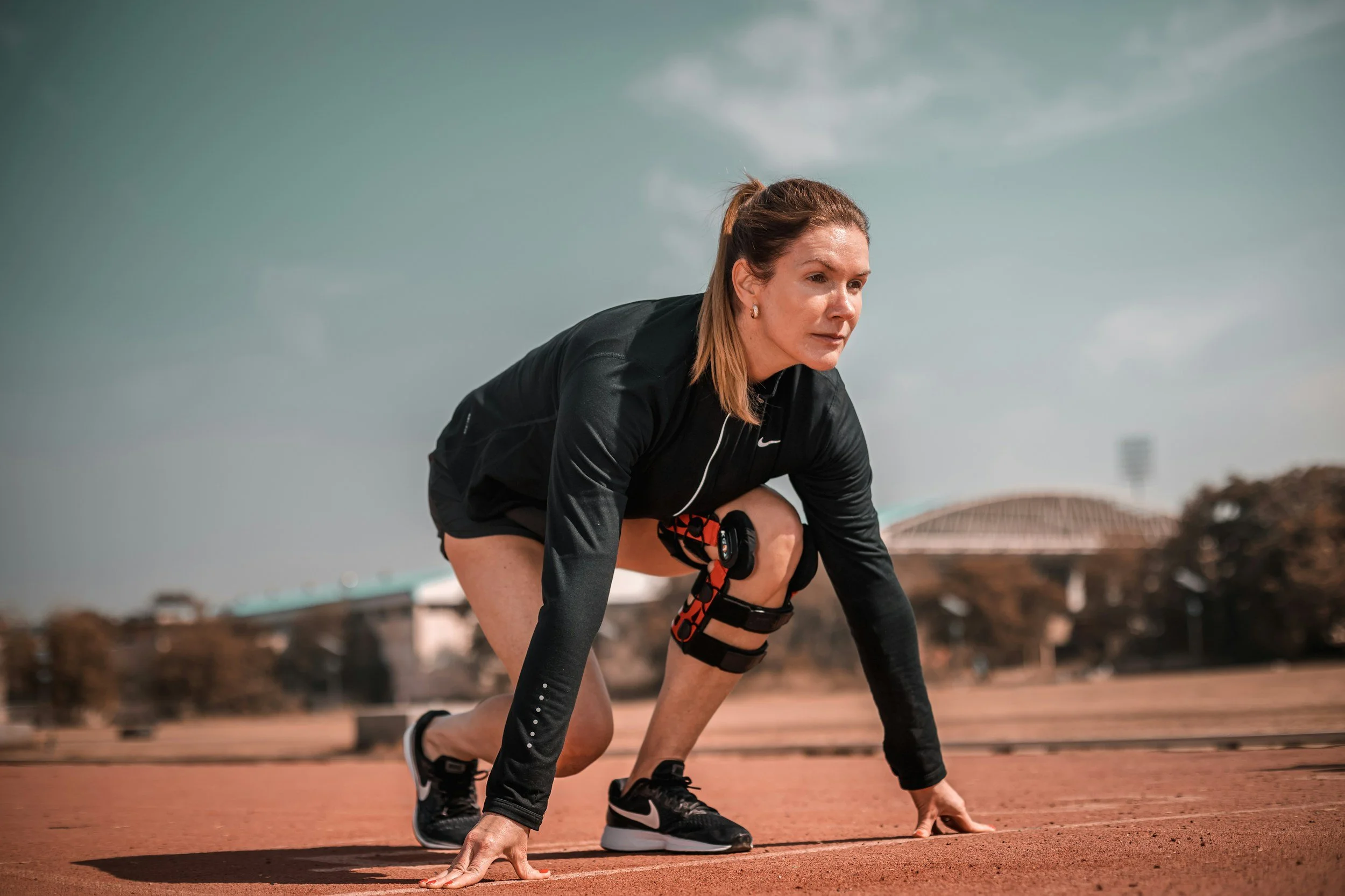 A woman in athletic gear, crouched in a starting position on a running track, preparing for a sprint. She is wearing black sportswear, running shoes, and a knee strap with a device, with an athletic stadium in the background.