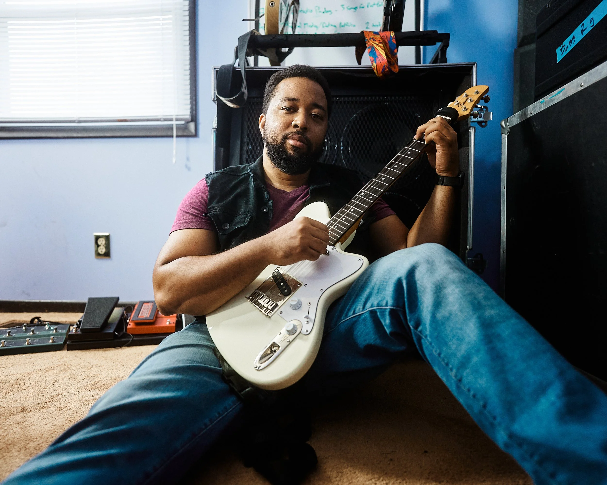 A man sitting on the floor in a music room holding a white electric guitar.