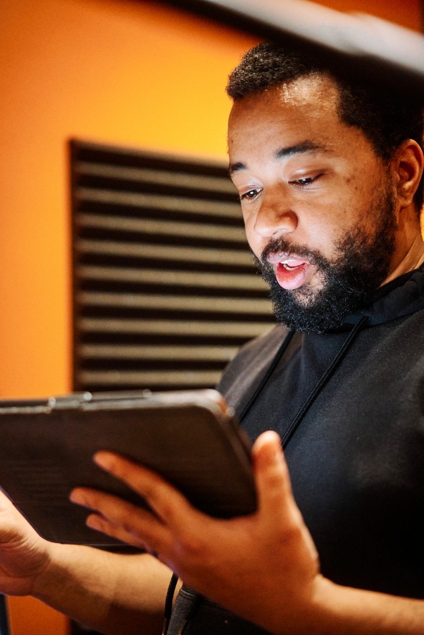 A man with a beard and short hair looking at a smartphone, wearing a black hoodie, with orange and black soundproofing panels in the background.
