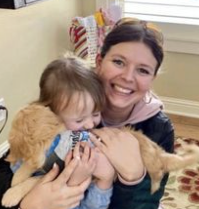 A woman smiling and hugging a young child and a small dog inside a home.