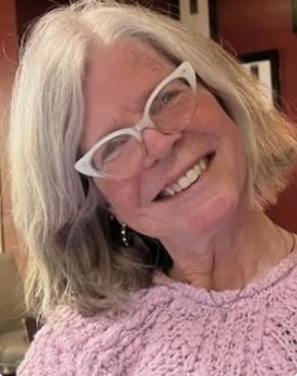 Smiling woman with gray hair, white glasses, and earrings, wearing a pink sweater.