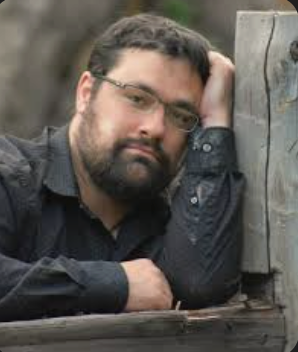Man with dark hair and beard, wearing glasses and a dark shirt, resting his arm on a wooden surface outdoors.