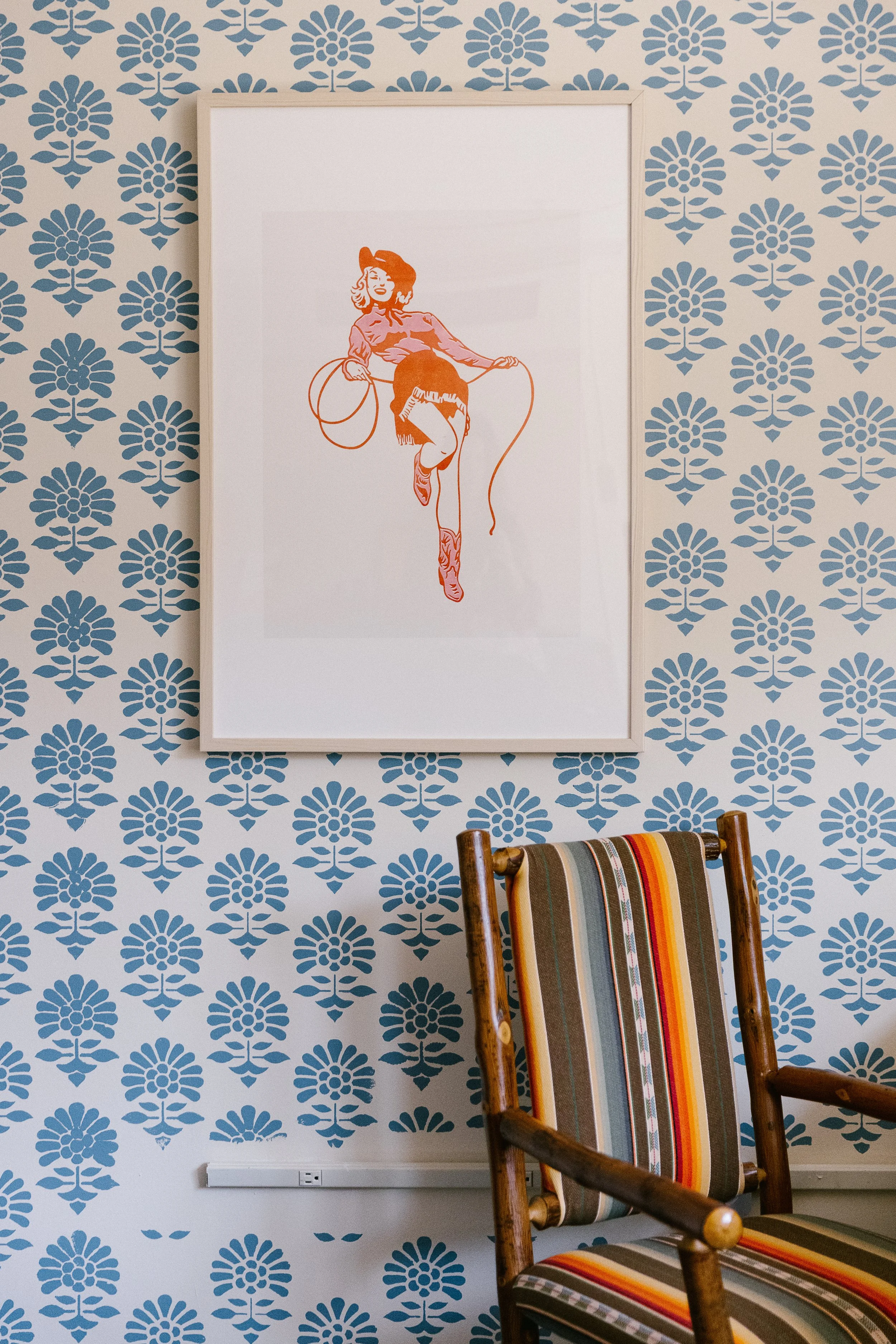 Decorative room with a blue floral patterned wallpaper, a framed artwork of a sketched woman with a rope, and a wooden rocking chair with a striped cushion.