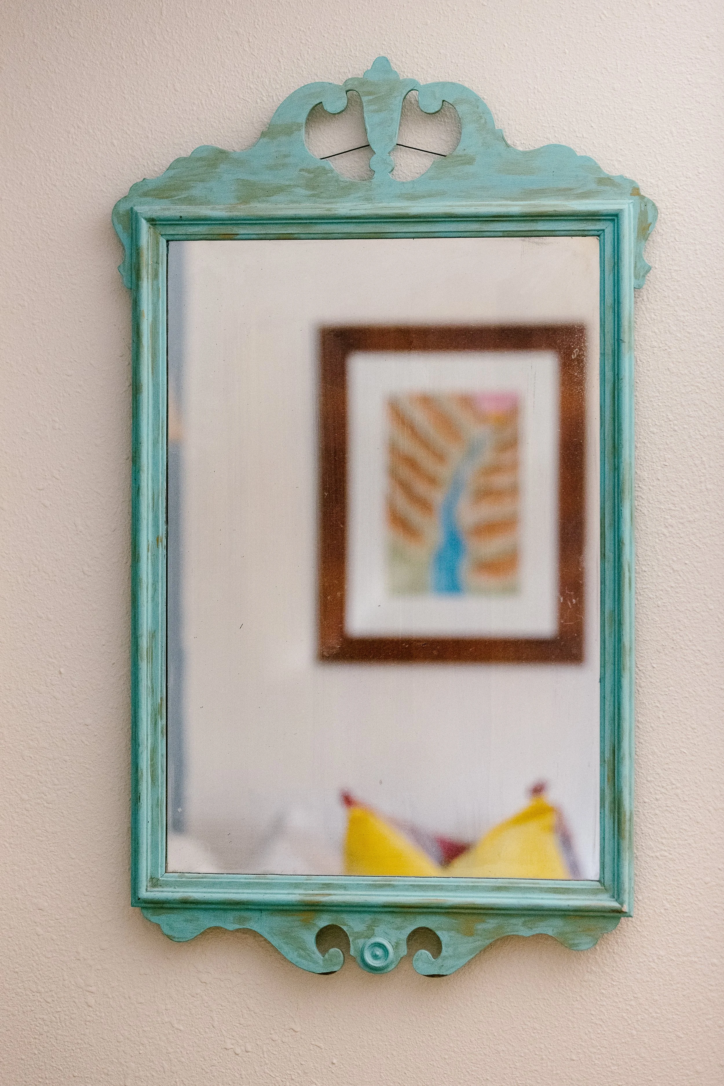 A decorative, vintage turquoise mirror hangs on a beige wall, reflecting a blurred framed artwork of a blue river and colorful landscape, with a yellow cushion or pillow at the bottom of the frame.