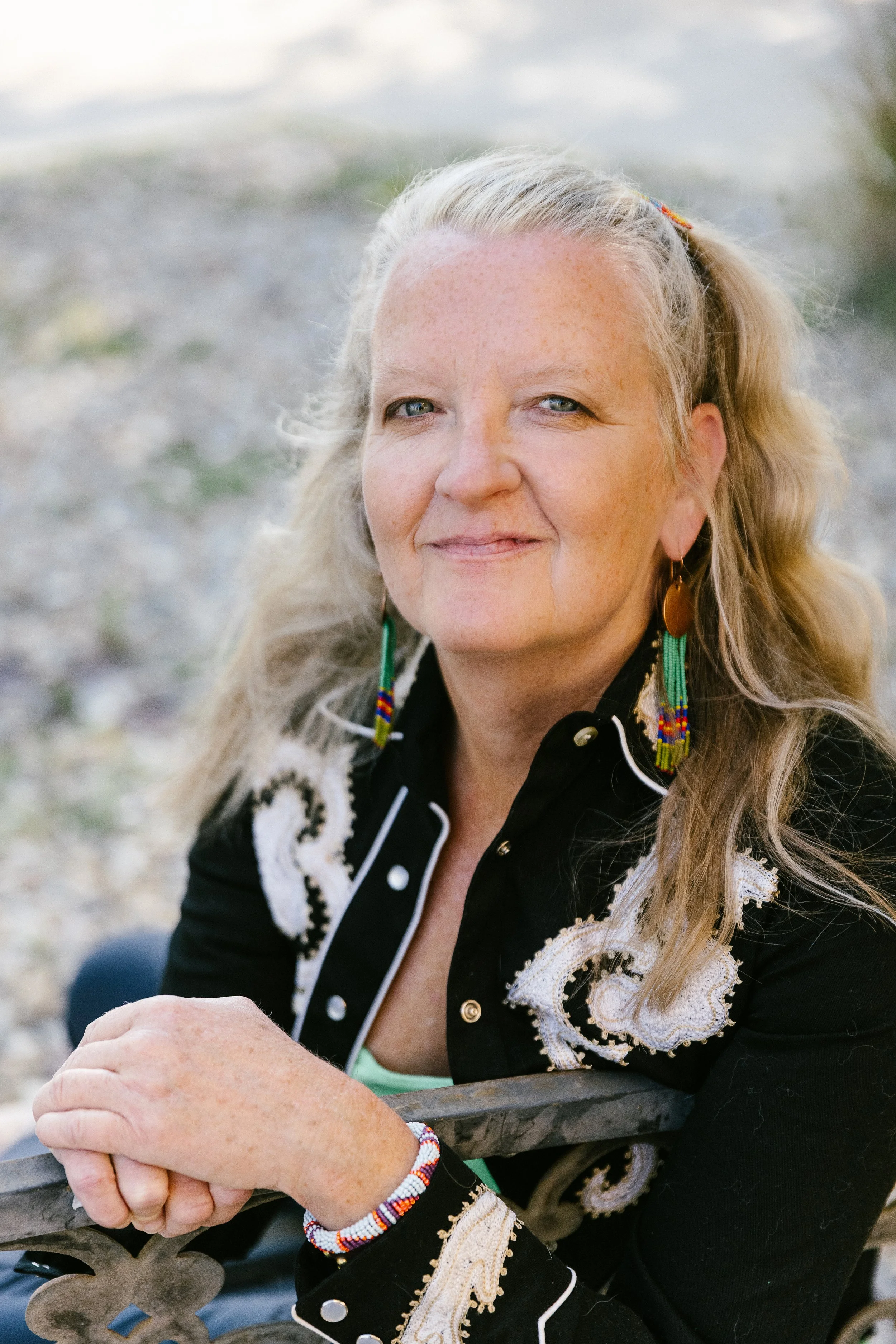 A woman with long blonde hair, blue eyes, and freckles, smiling outdoors, wearing a black embroidered jacket, colorful beaded earrings, and a beaded bracelet, sitting and holding onto a metal rail.