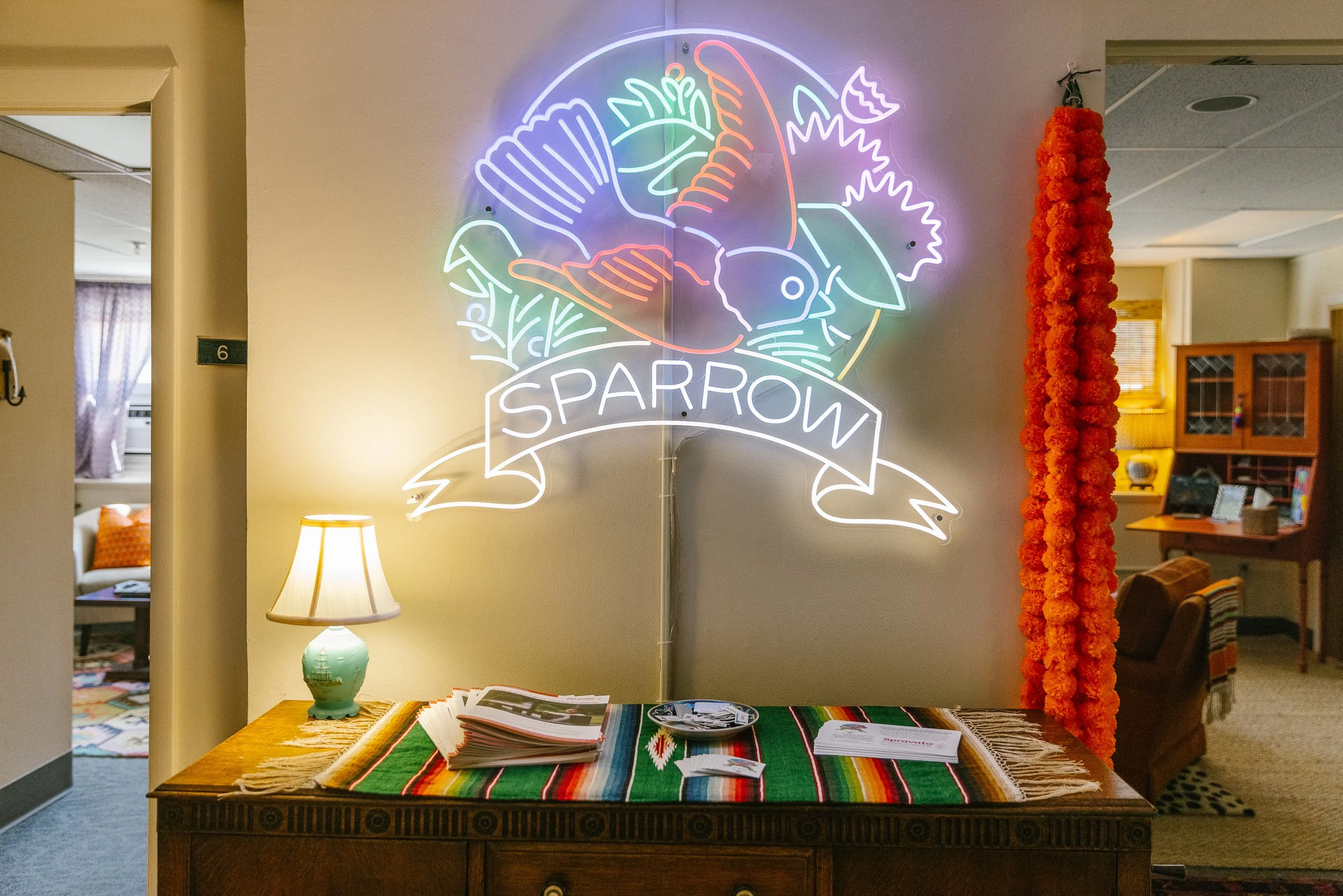 Neon sign of a bird surrounded by leaves with the word 'Sparrow' on a ribbon banner, mounted on a wall in a cozy room.
