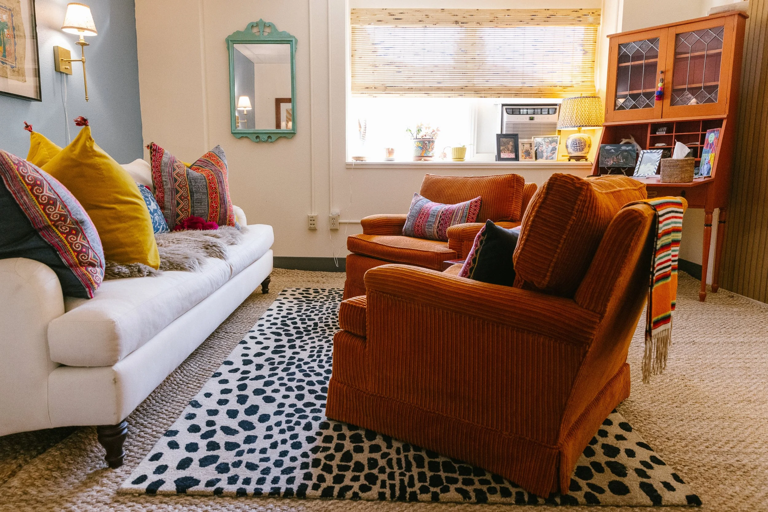 Living room with a white sofa, orange and brown armchairs, a black and white patterned rug, and various colorful throw pillows. There is a window with blinds, a cabinet with display items, and framed photos on the windowsill.