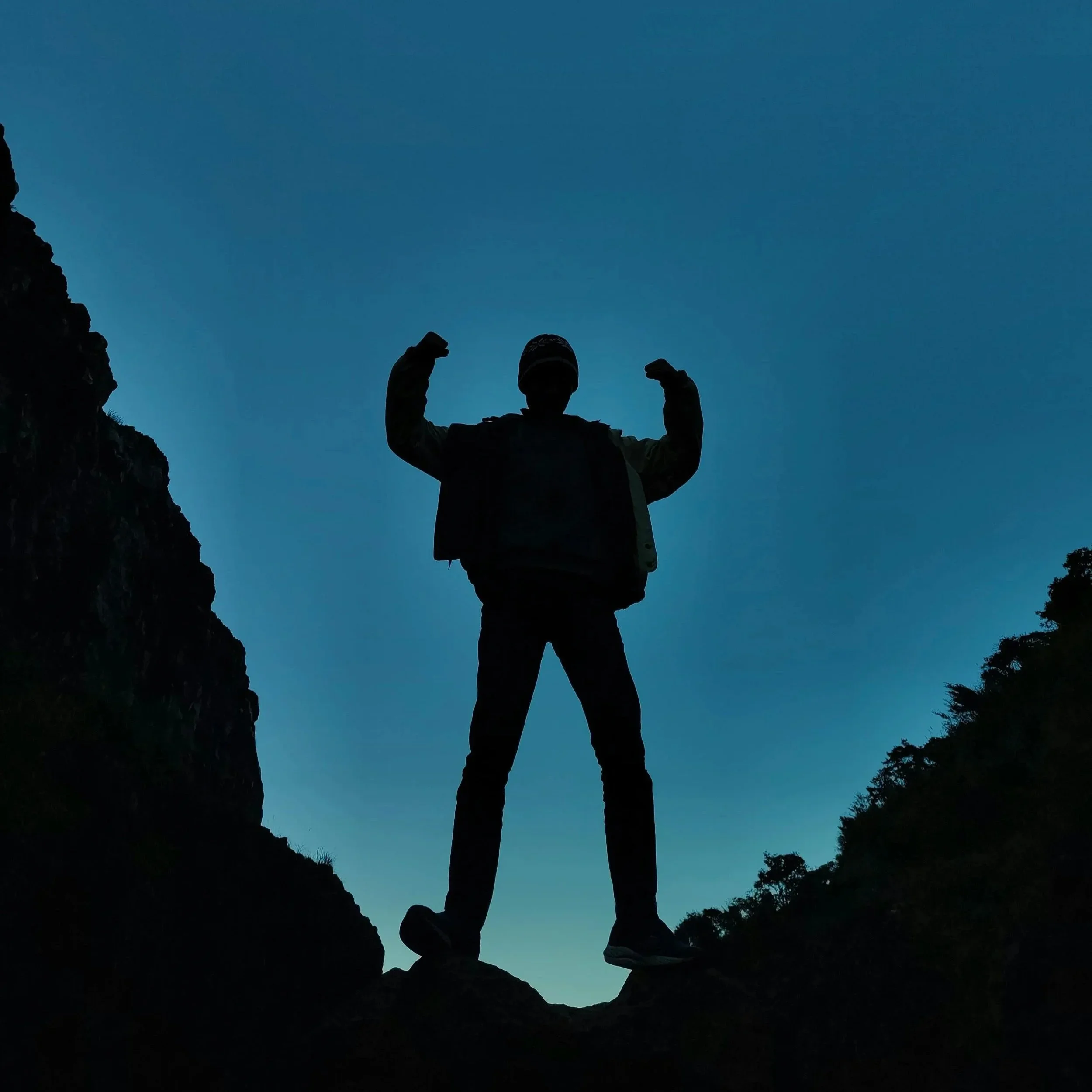 Silhouette of a person standing on rocks between two cliffs with a blue sky in the background, raising their arms.