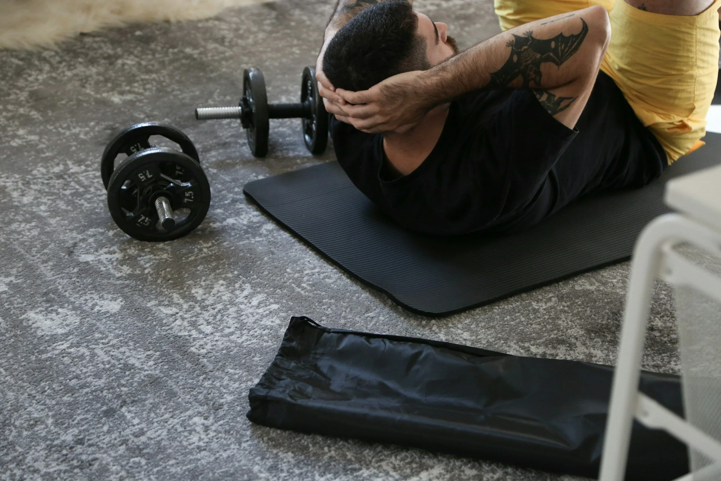 A man lying on a black exercise mat doing sit-ups, with a pair of dumbbells nearby, in a gym or workout area.