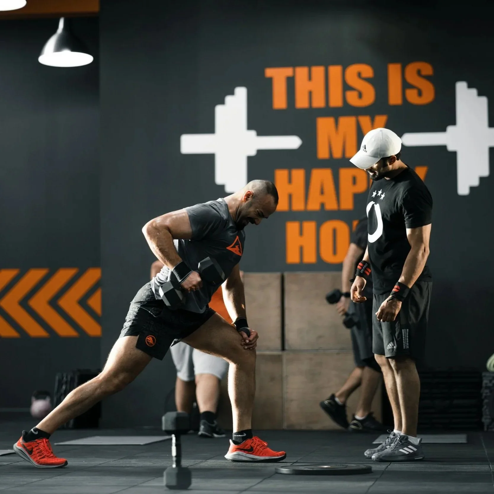 Men working out in a gym with a motivational sign on the wall that reads 'This is my happy hour'