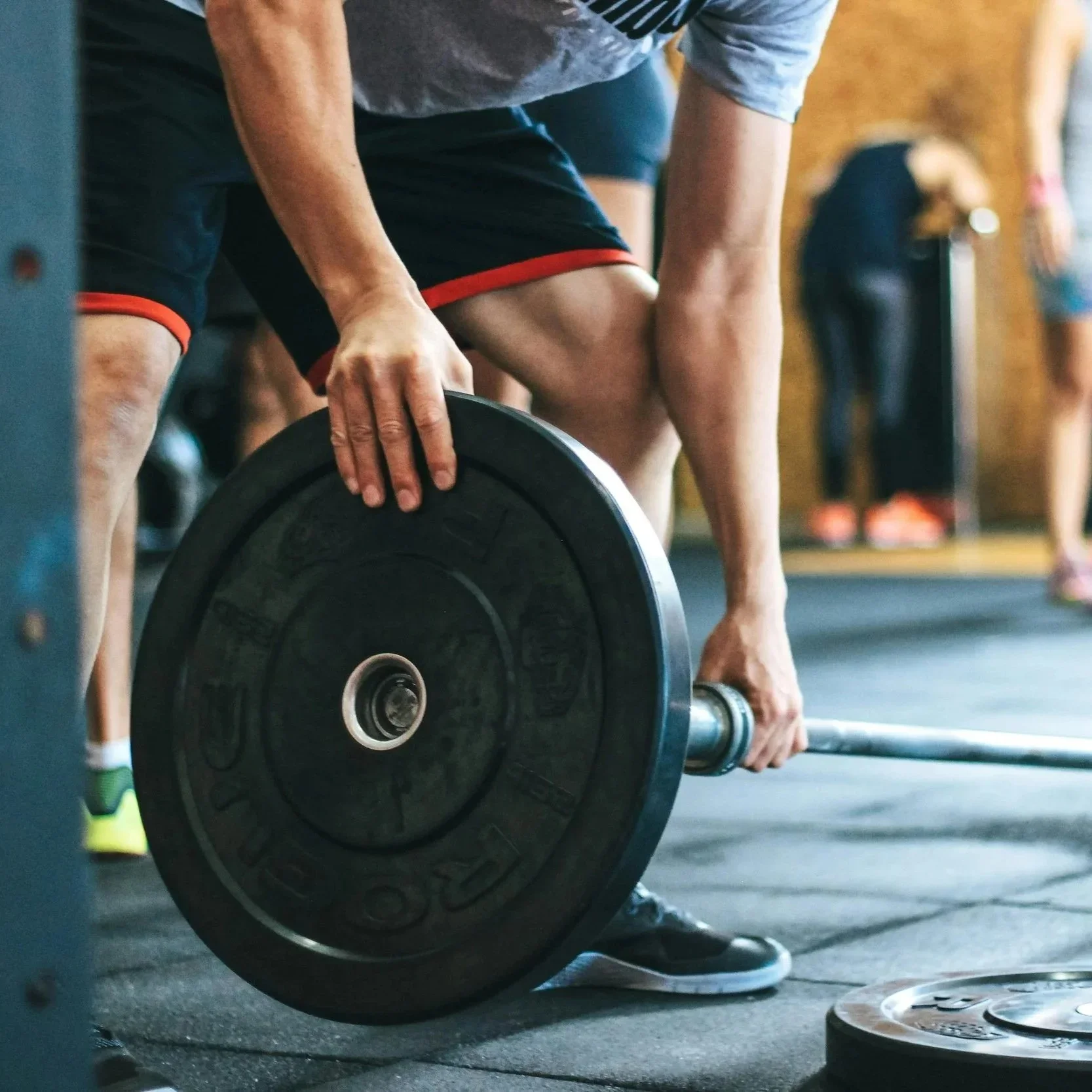 A person is lifting a barbell during a workout in a gym, wearing a grey t-shirt and black shorts with red trim. The focus is on their hands gripping the barbell, with a weight plate on the barbell, and other gym equipment visible in the background.