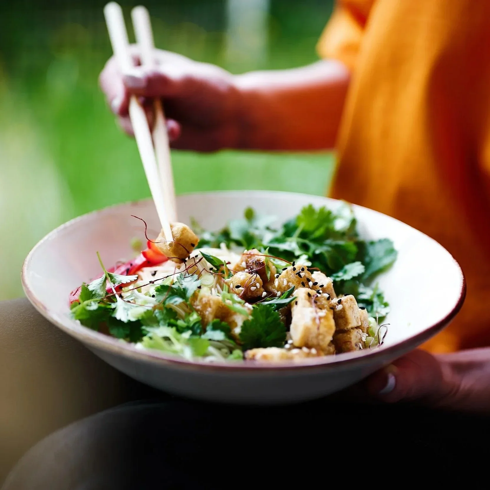 A hand holding a bowl of Asian-style salad with greens, tofu, and sesame seeds, as chopsticks pick up a piece of tofu.