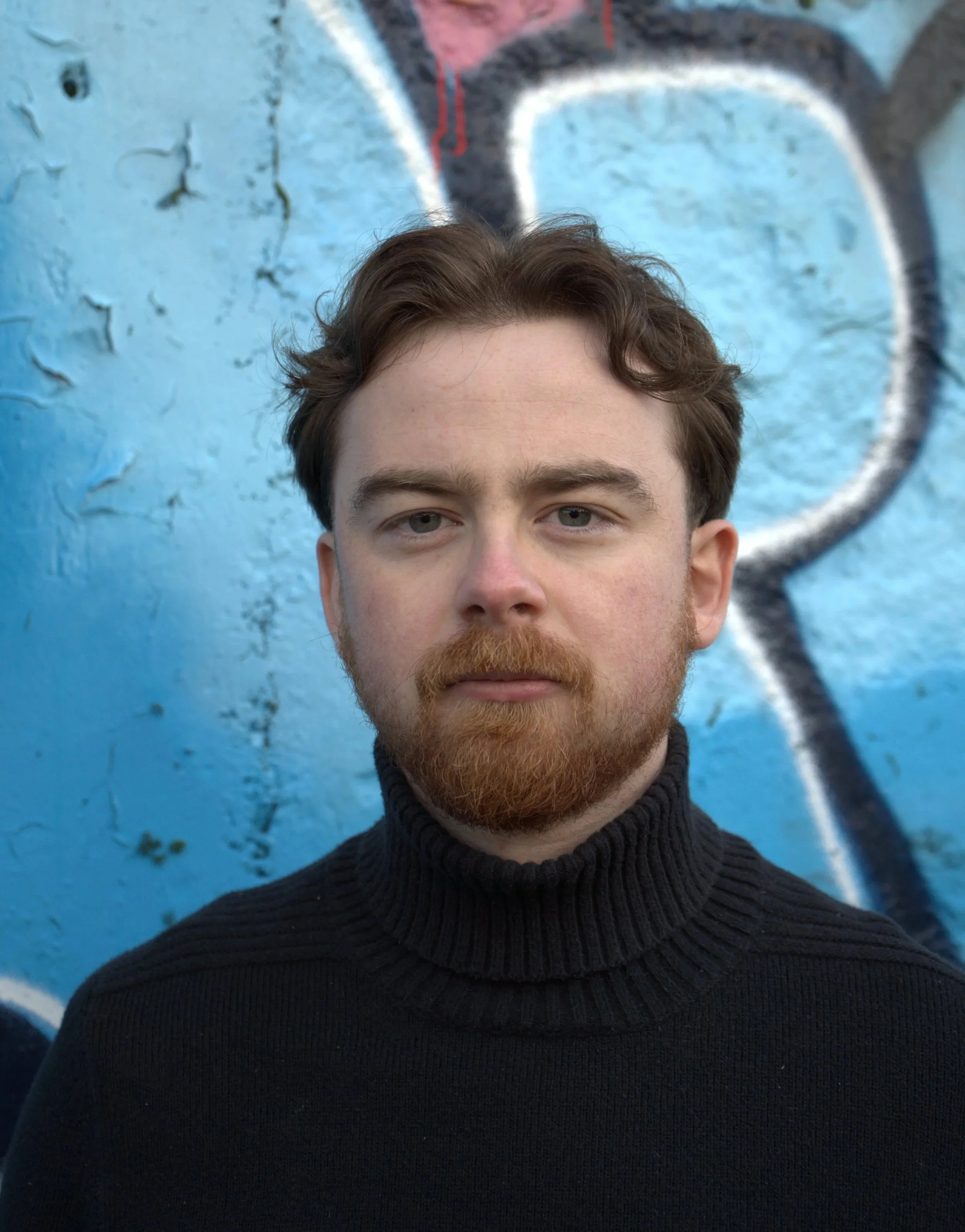 A young man with red hair and a beard wearing a black turtleneck sweater standing in front of a blue graffiti wall.