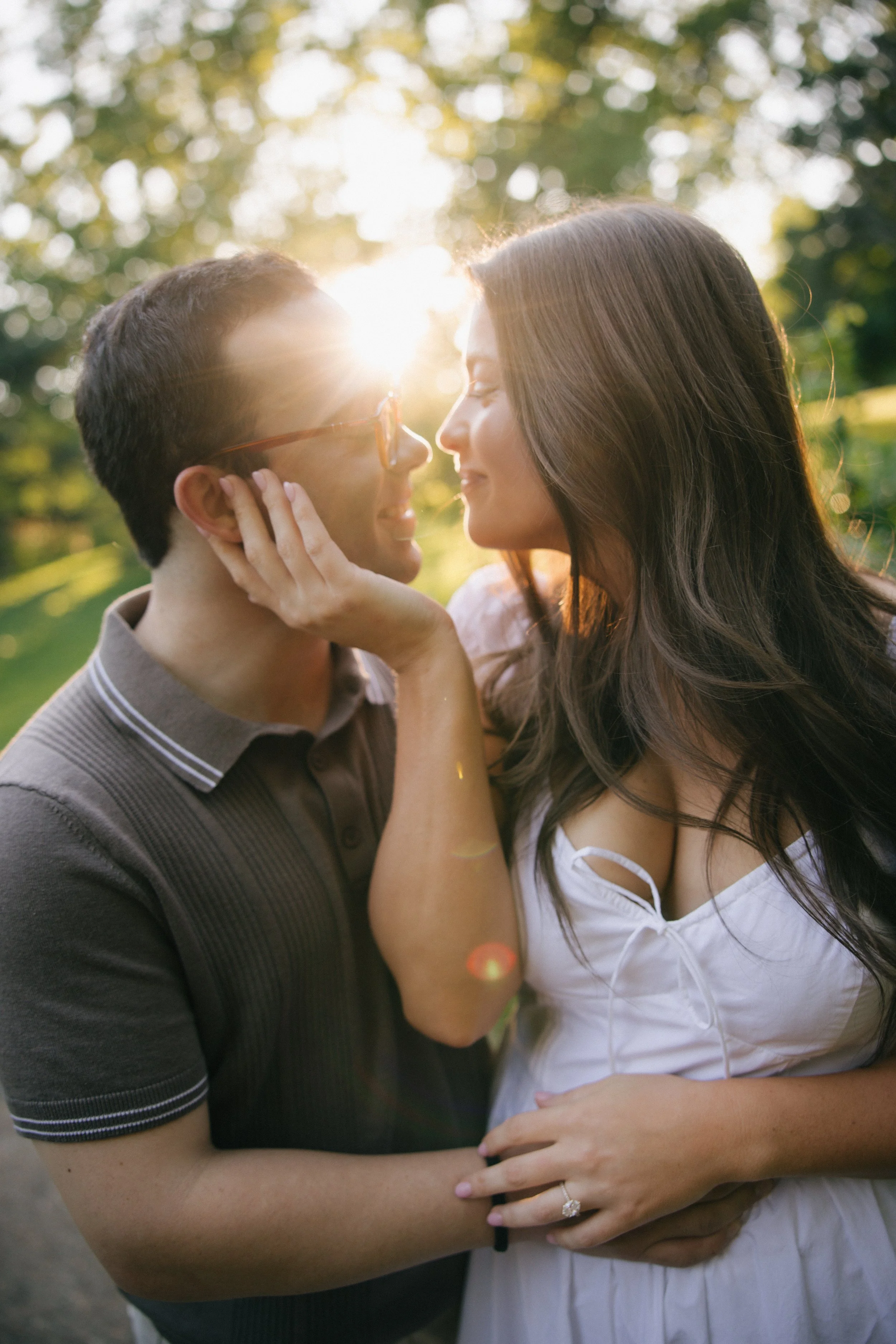 A couple sharing an intimate moment outdoors during sunset, with their noses nearly touching and smiling at each other. The woman is gently holding the man's face.