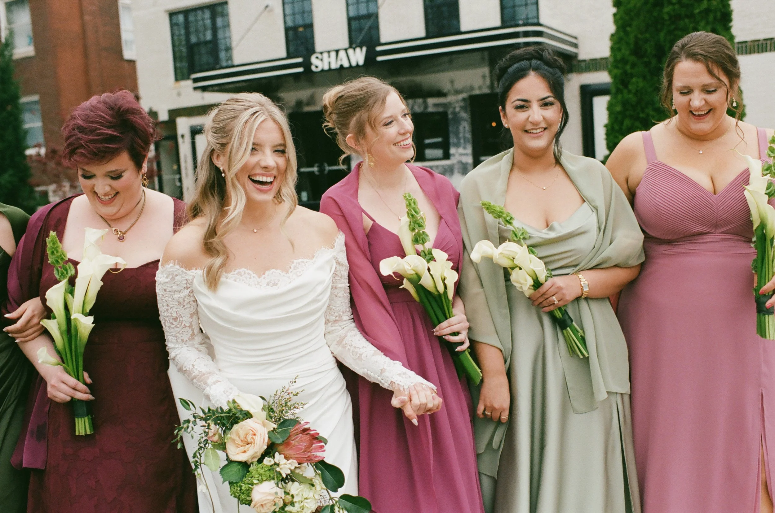 A group of women dressed in colorful dresses, holding flowers, standing together outside, smiling and laughing during what appears to be a celebration or wedding.