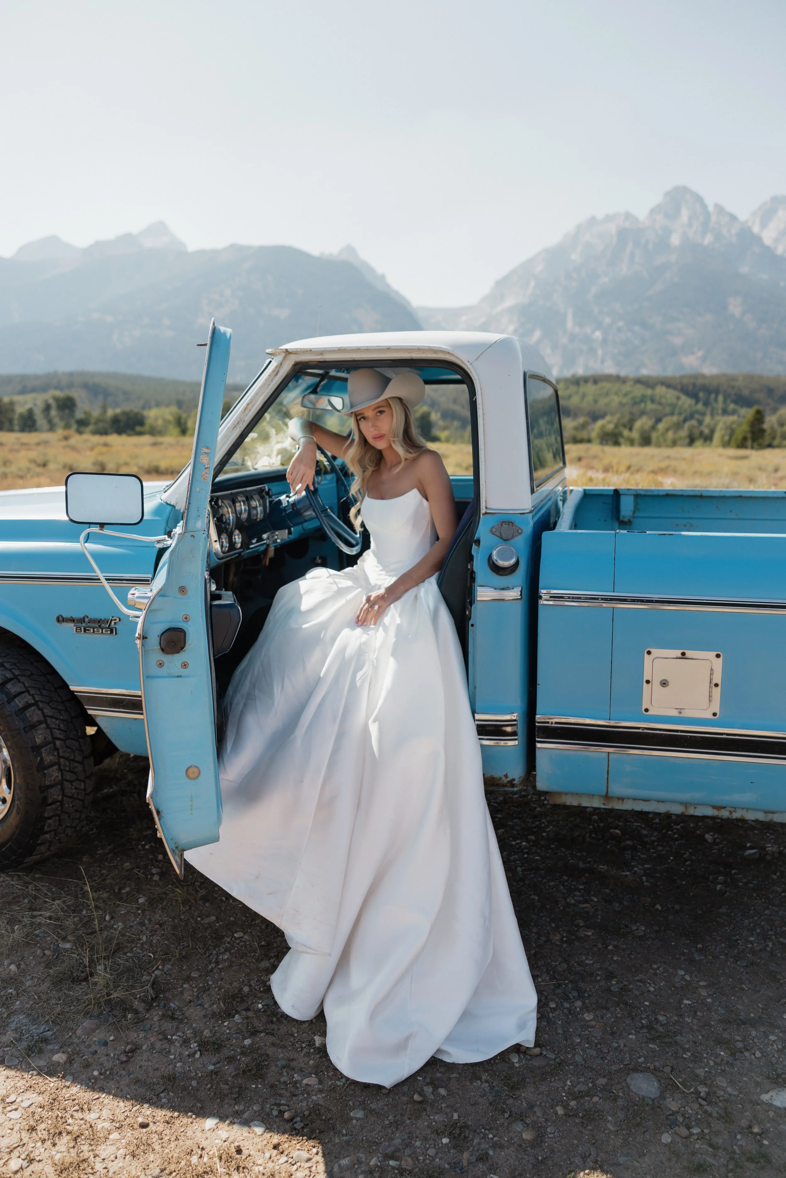 Woman in a white dress and cowboy hat sitting in the driver seat of a blue vintage pickup truck with mountains in the background.