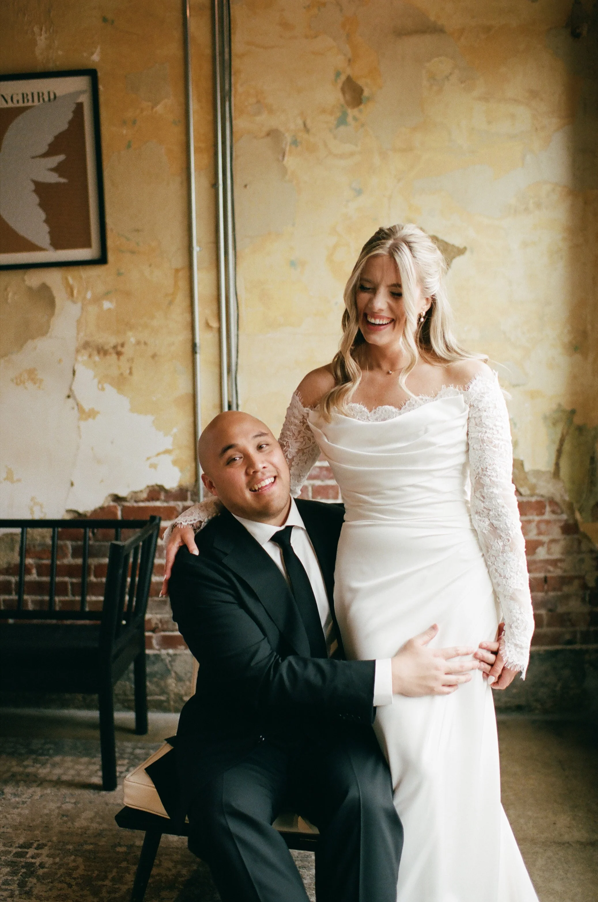 A woman in a wedding dress and a man in a tuxedo sitting and standing in front of a brick wall with peeling paint, smiling and posing for the camera.