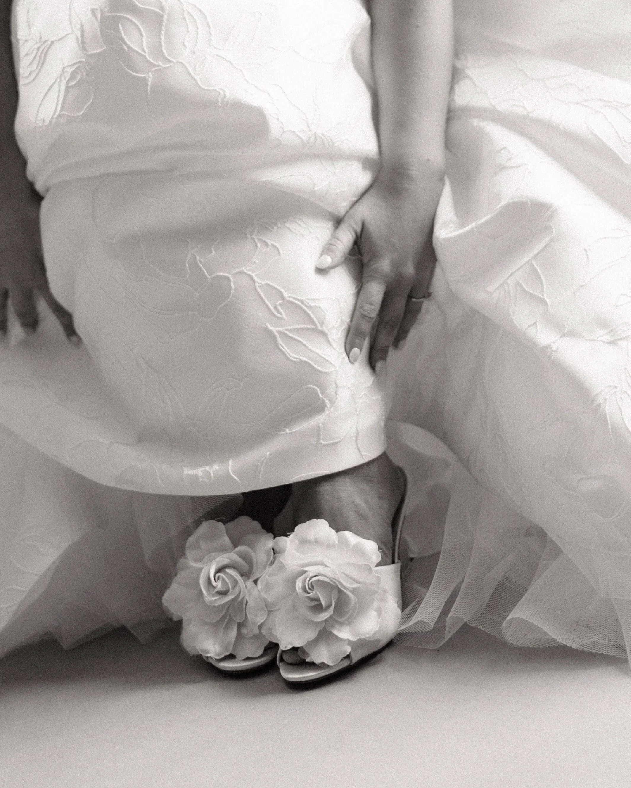 Close-up black and white photo of a wedding ring on a woman's finger, resting on a fabric with a floral pattern.