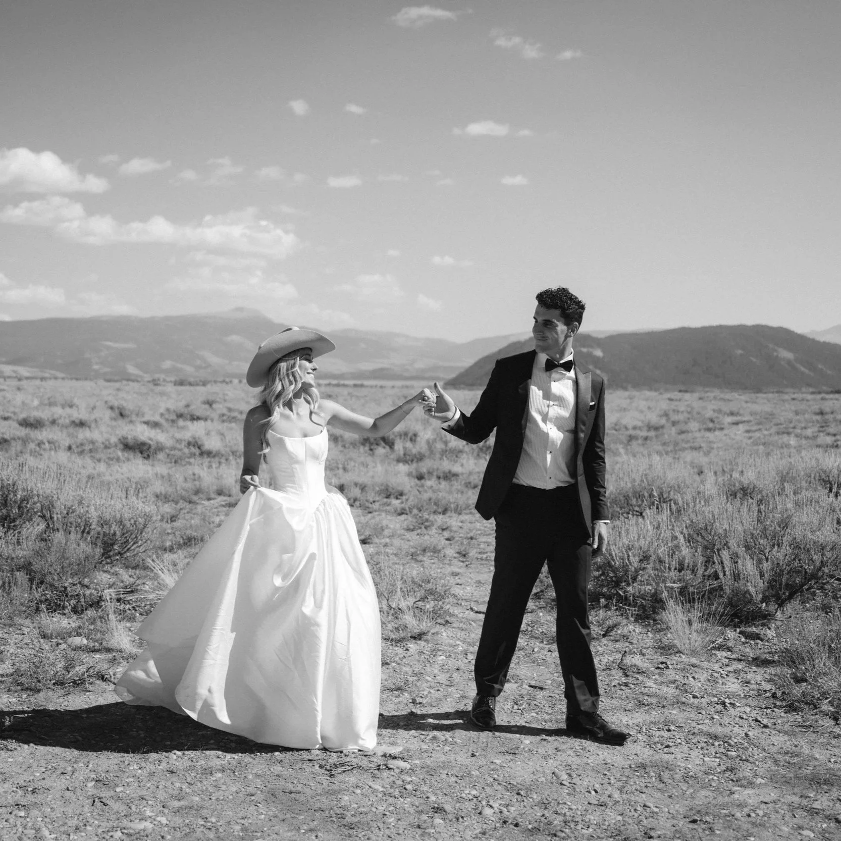 A black and white photo of a bride and groom in a desert landscape, holding hands. The bride is wearing a white strapless wedding dress and a cowboy hat, smiling. The groom is in a tuxedo with a bow tie, looking at her, smiling.
