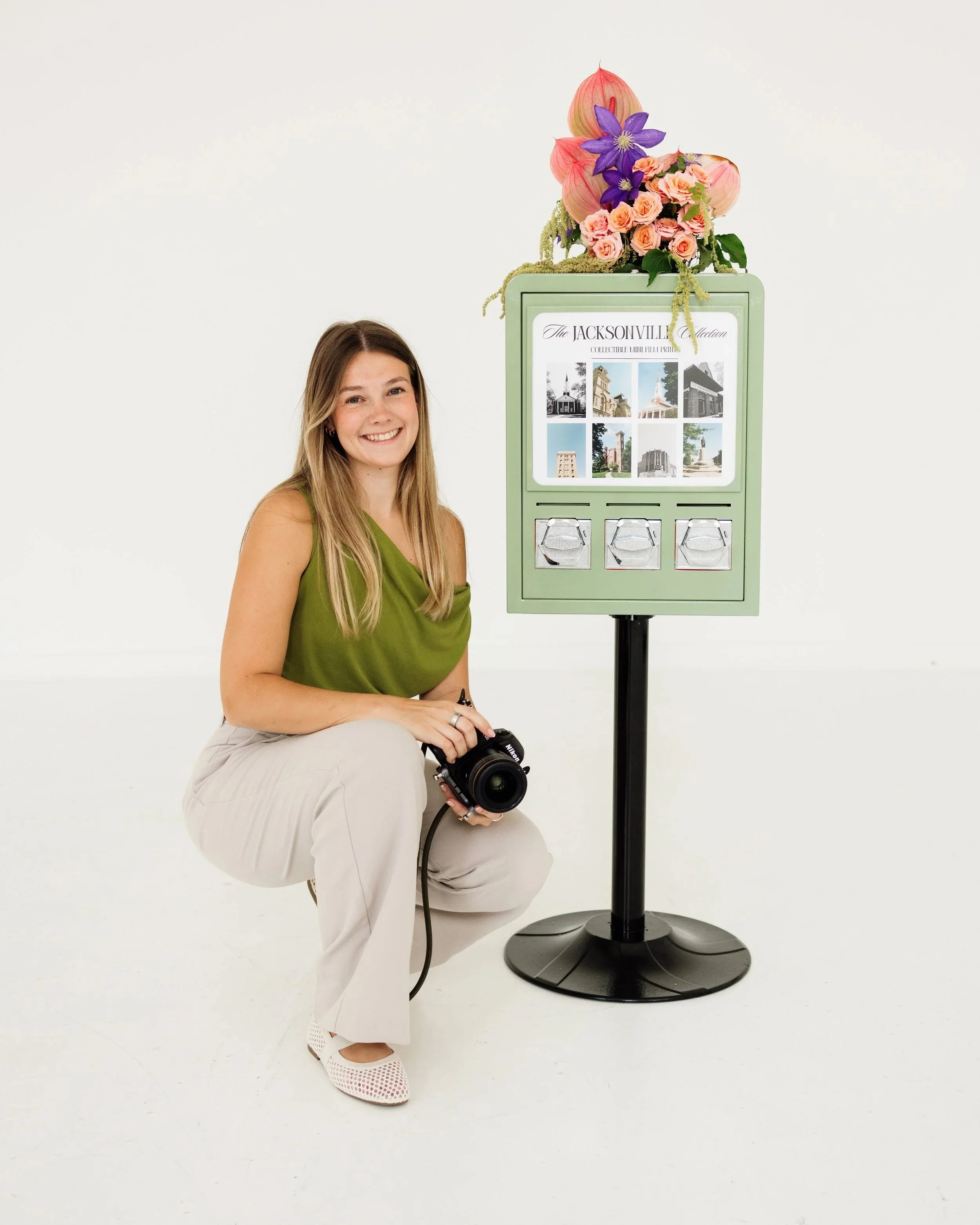 A young woman with long blonde hair, wearing a green sleeveless top and light-colored pants, is kneeling with one knee on the ground, holding a camera, next to a display stand with a floral arrangement and a photo display, against a plain white background.
