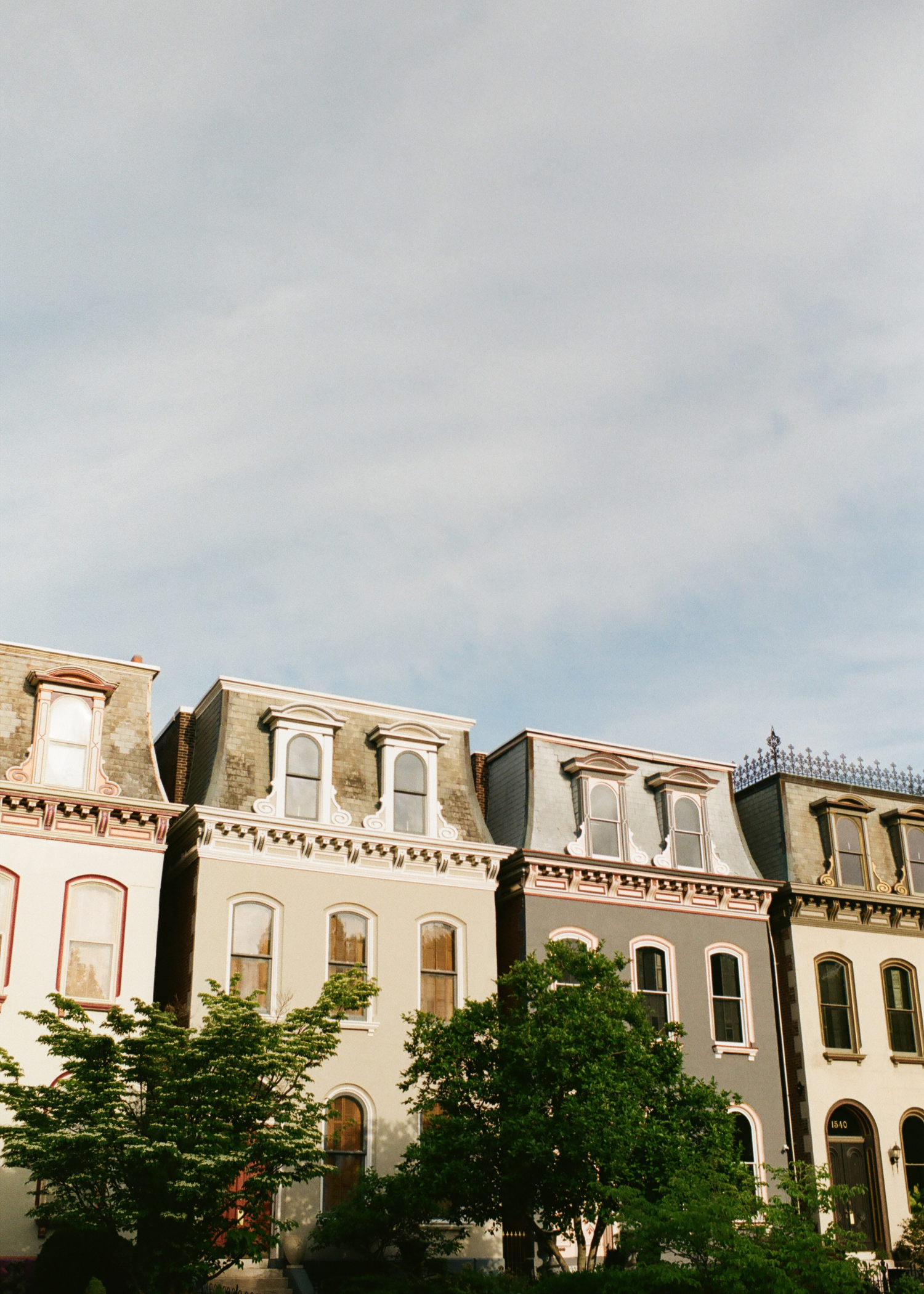 A row of colorful Victorian-style houses with ornate architectural details, multiple stories, and large windows, with green trees in the foreground and a cloudy sky above.