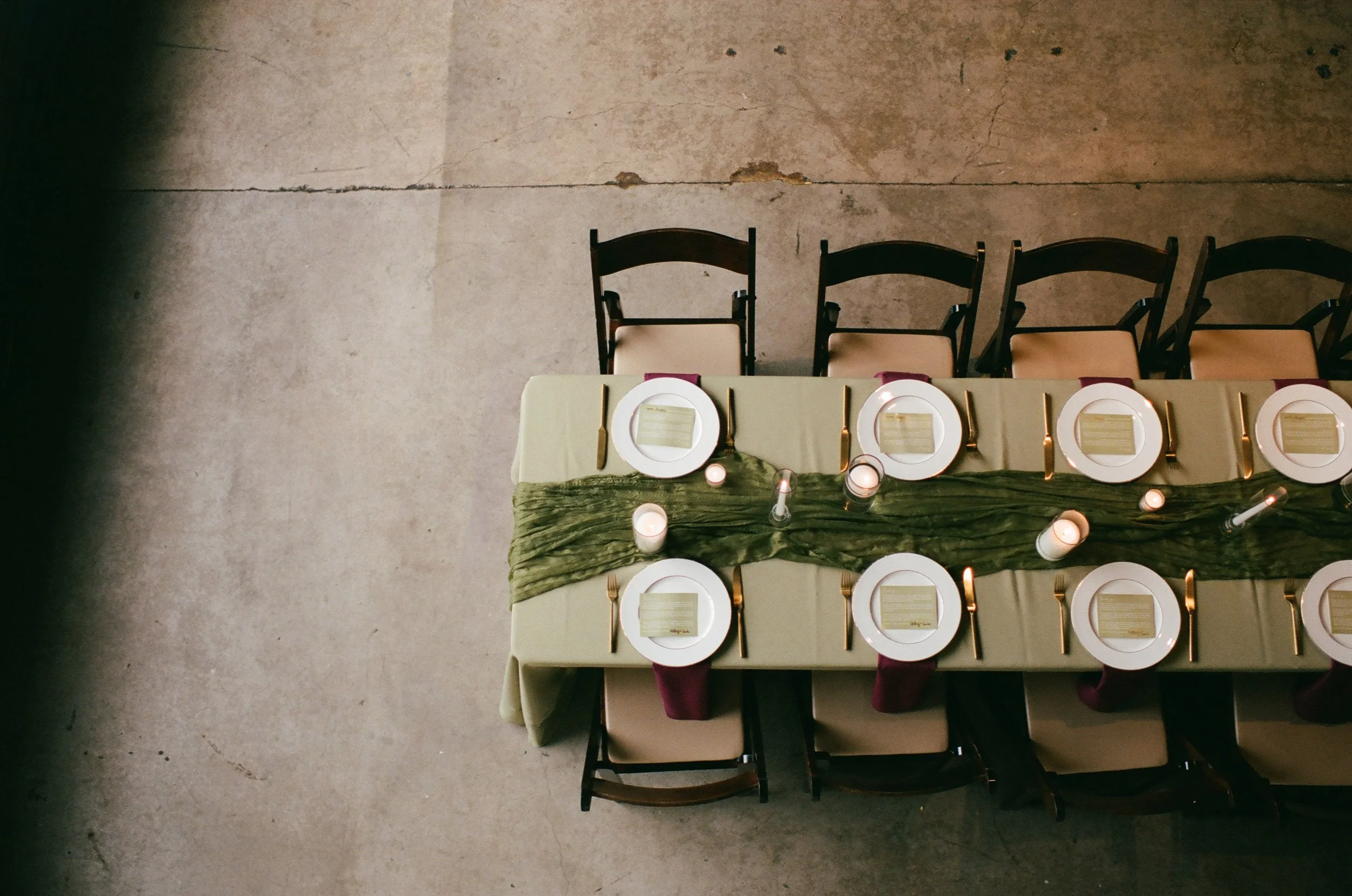 A long banquet table set for a meal with plates, napkins, and gold utensils, decorated with a green runner and candles, with chairs arranged on either side against a concrete floor.