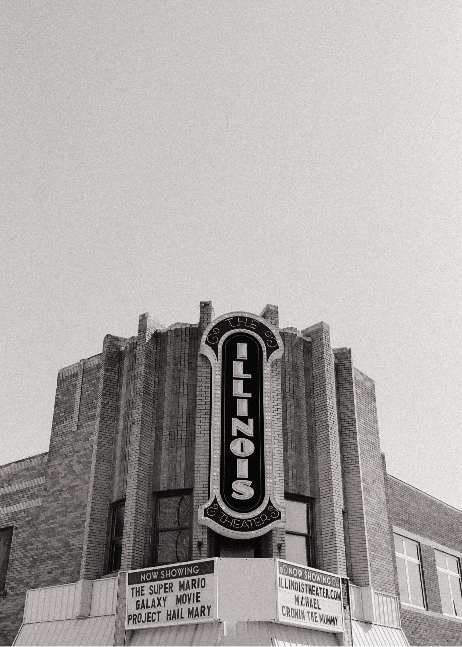 A black and white photograph of a brick theater building with a vertical sign that says "THE ILLINOIS THEATER." There is a marquee at the bottom with text advertising movies, including "THE SUPER MARIO GALAXY MOVIE" and "PROJECT HAIL MARY."