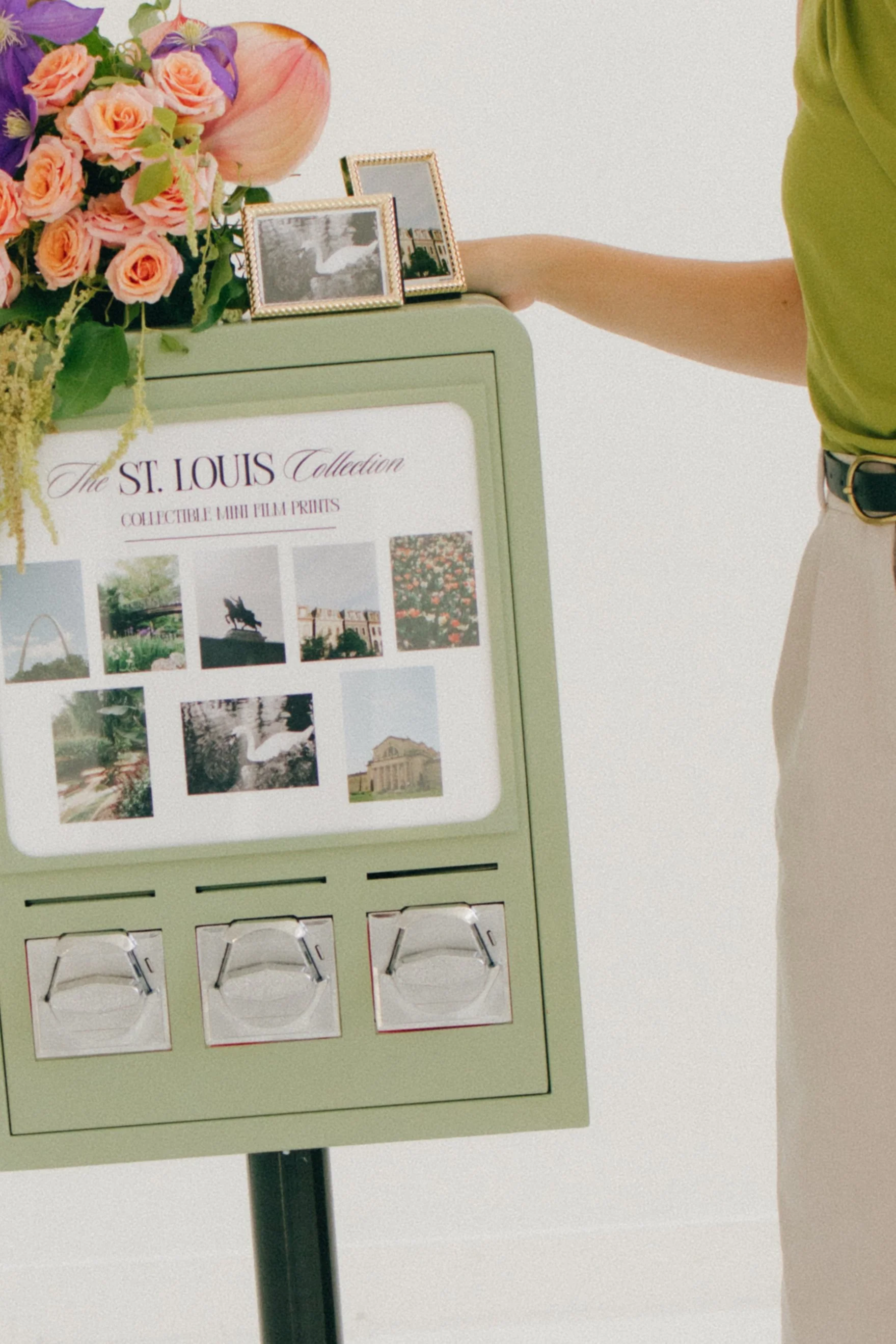 A display board titled 'The St. Louis Collection' features images of landmarks and scenes from St. Louis, Missouri, including the Gateway Arch and other cityscapes. A person in a green shirt and beige pants reaches out towards the board, which is topped with a floral arrangement and framed photographs.