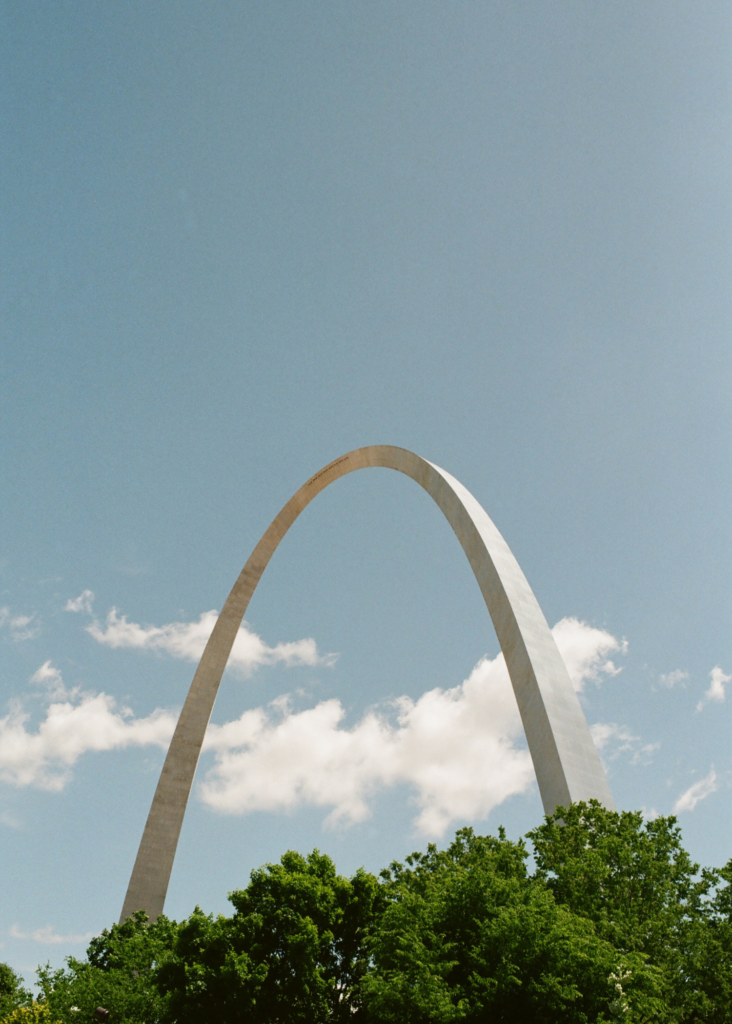 View of the Gateway Arch in St. Louis, Missouri, with a partly cloudy blue sky and green trees in the foreground.