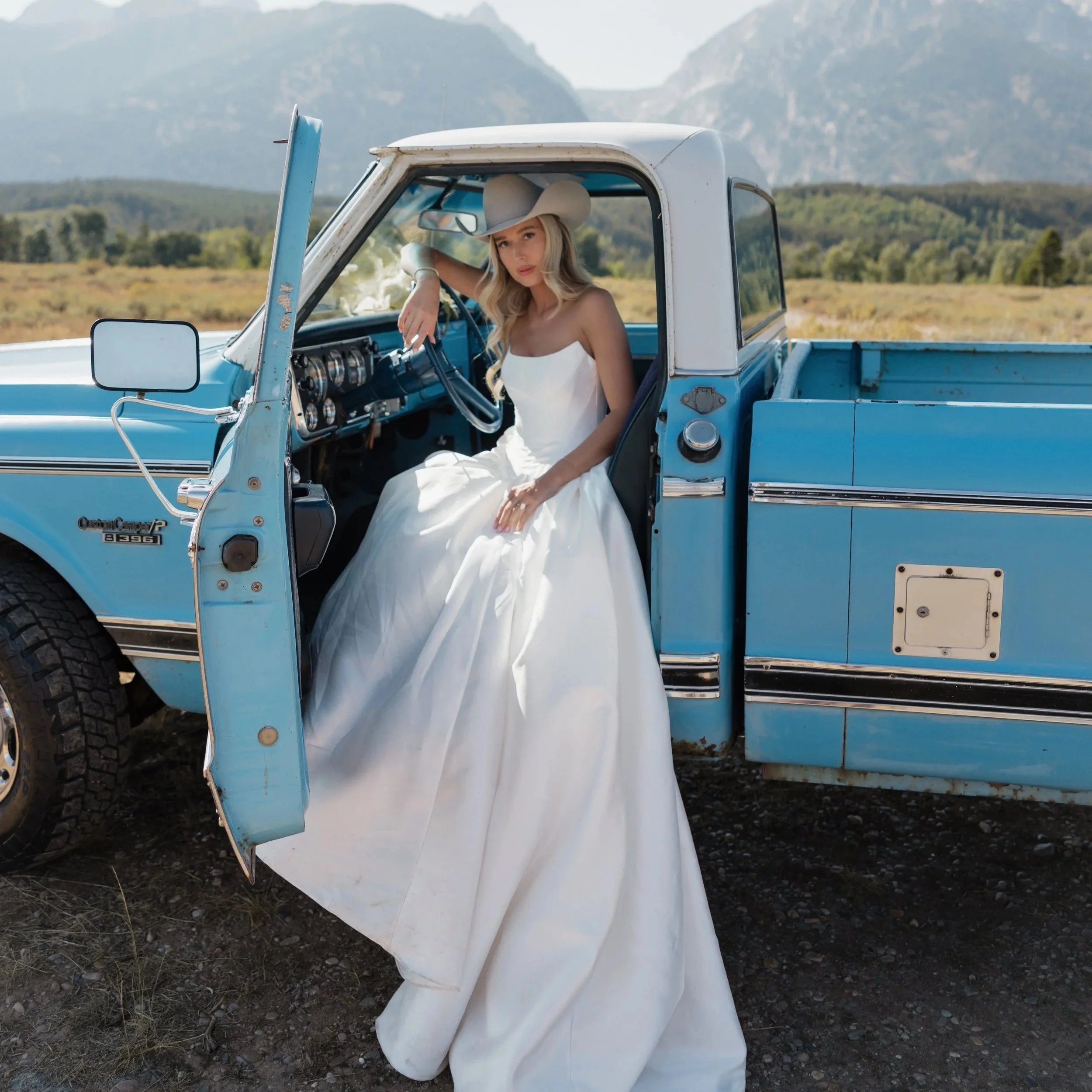 A woman in a white dress and cowboy hat sitting in a vintage blue pickup truck on a rural landscape with mountains in the background.