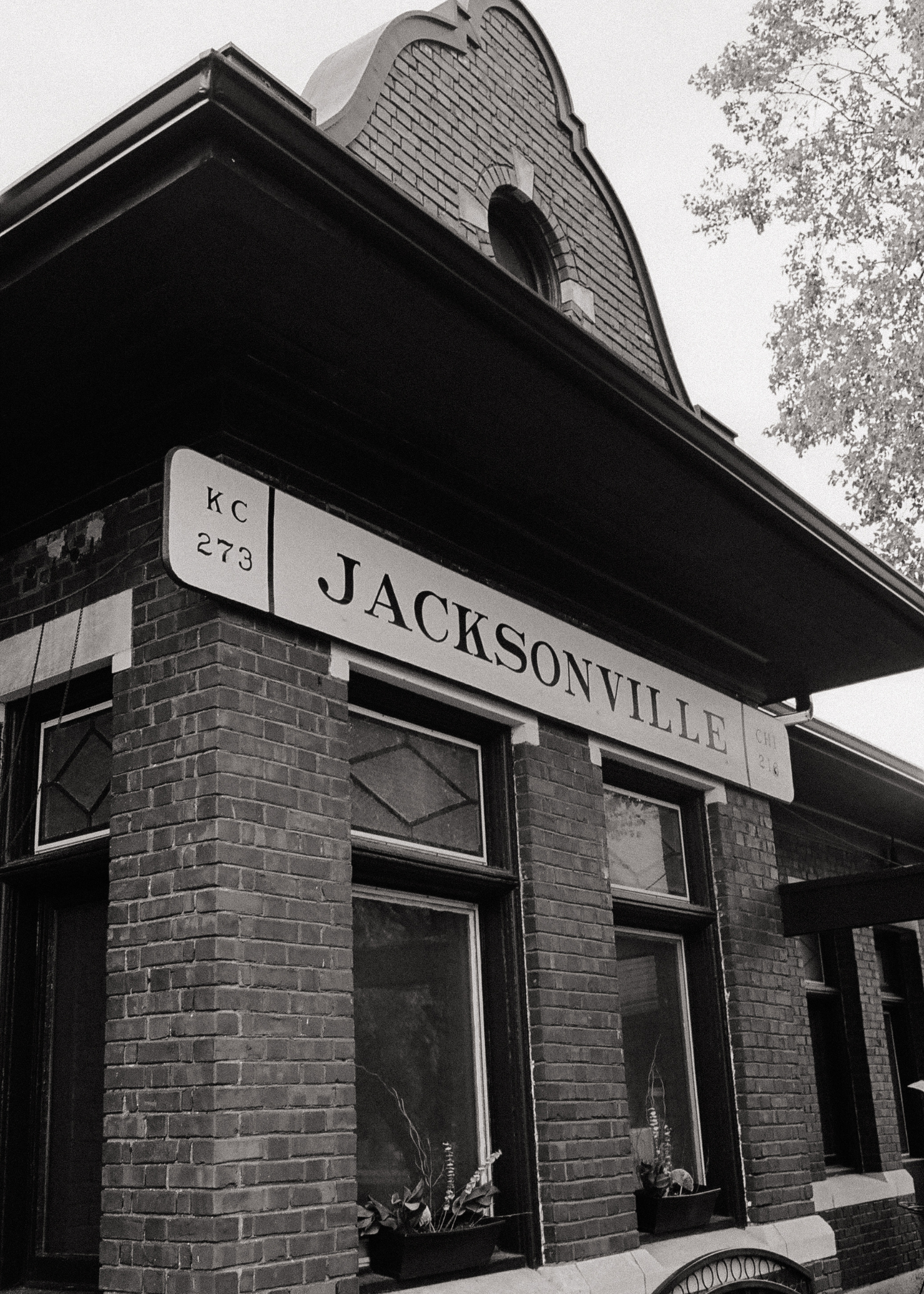 Black and white photo of a corner of a brick building with a sign that says 'Jacksonville' and a smaller sign with 'KC 273'. The building has large windows with flower pots underneath.