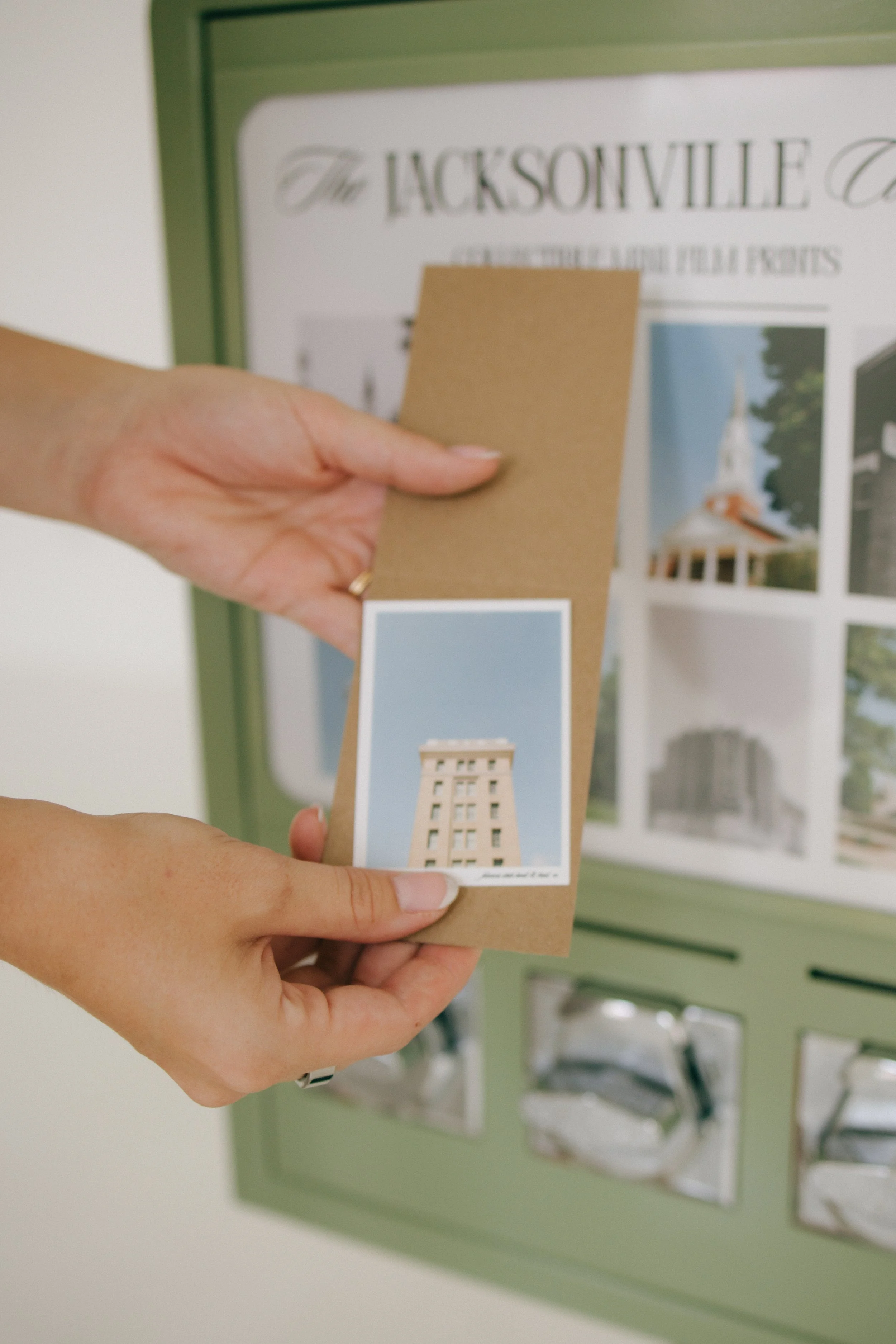 Person holding a small postcard with an image of a tall building, in front of a display board with posters of similar buildings.